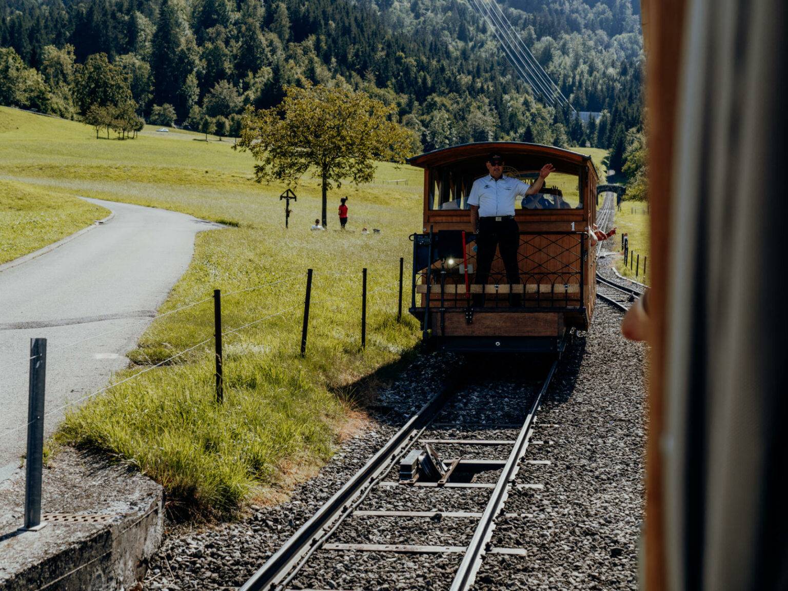 Eine Oldtimer-Standseilbahn fährt über Gleise auf Kies, im Hintergrund liegt ein Wald, links neben der Bahn verläuft ein Weg.