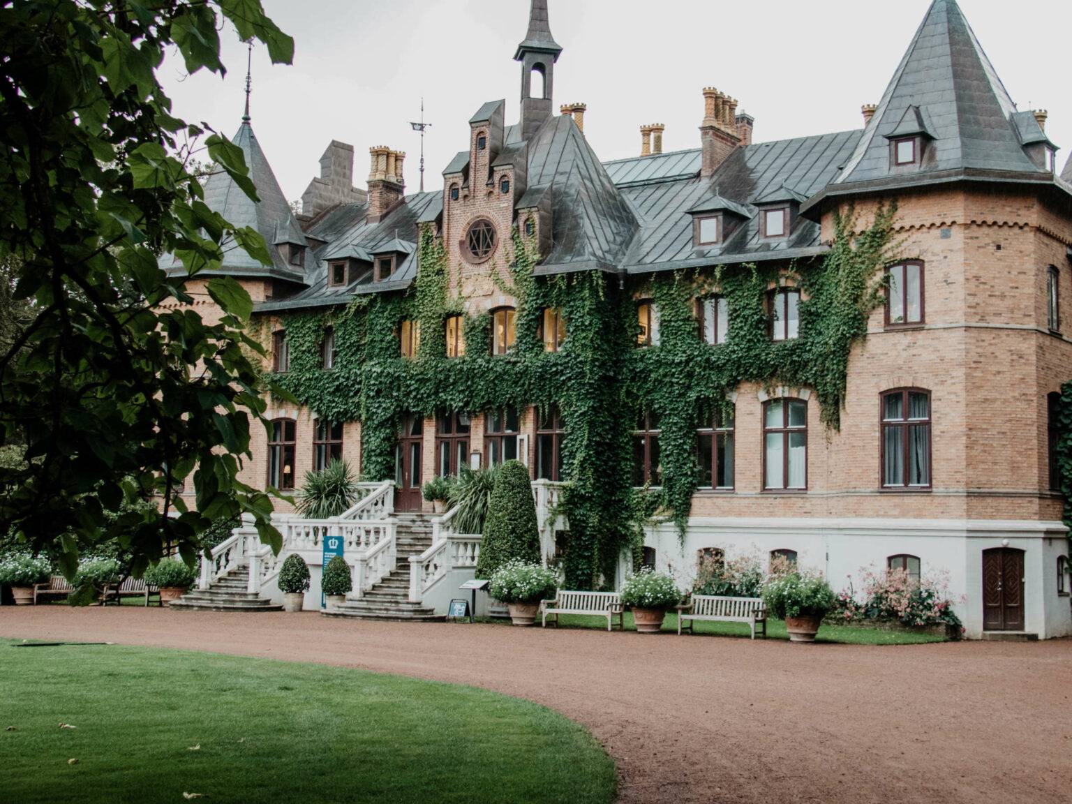 Das Schloss Sofiero liegt in einer grünen Parkanlage hinter einem roten Steinweg, das herrschaftliche Anwesen ist mit Efeu bewachsen und hat ein graues Dach, zwei Erker stehen hervor.
