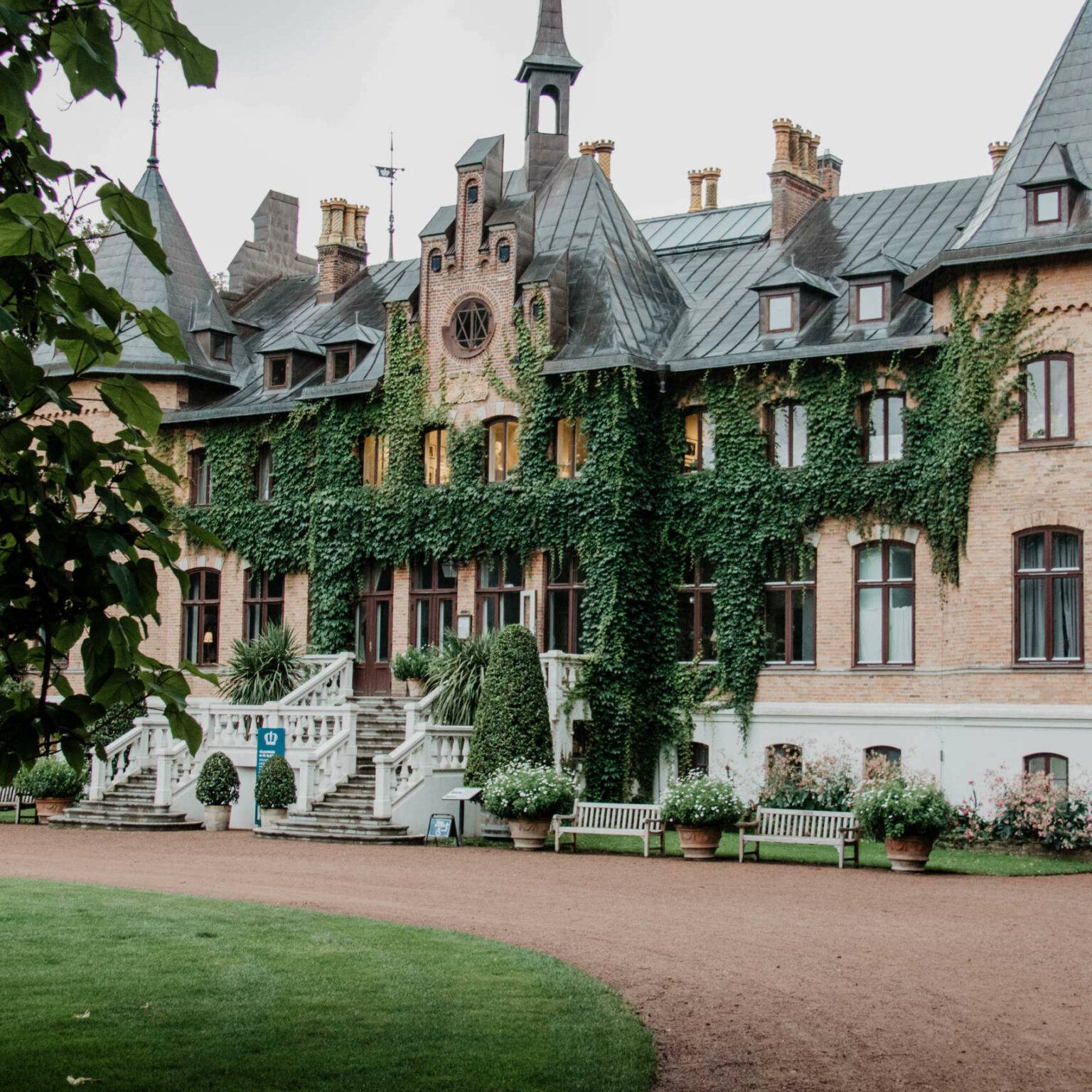 Das Schloss Sofiero liegt in einer grünen Parkanlage hinter einem roten Steinweg, das herrschaftliche Anwesen ist mit Efeu bewachsen und hat ein graues Dach, zwei Erker stehen hervor.