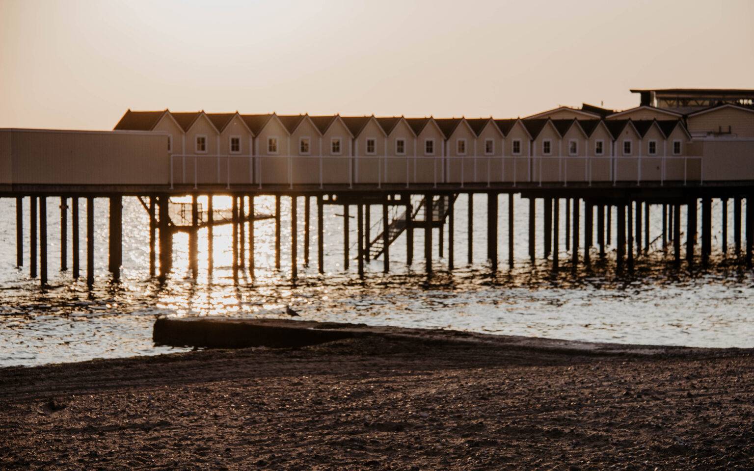 Eine lange Reihe von Häusern auf Stelzen, in denen sich Kaltbadehäuser befinden, liegen ein hohes Stück über dem seichten Wasser in der Abenddämmuerng, der Sand vom Strand färbt sich dunkel im Licht, eine Treppe aus dem Wasser führt die weißen Häuser hinauf.