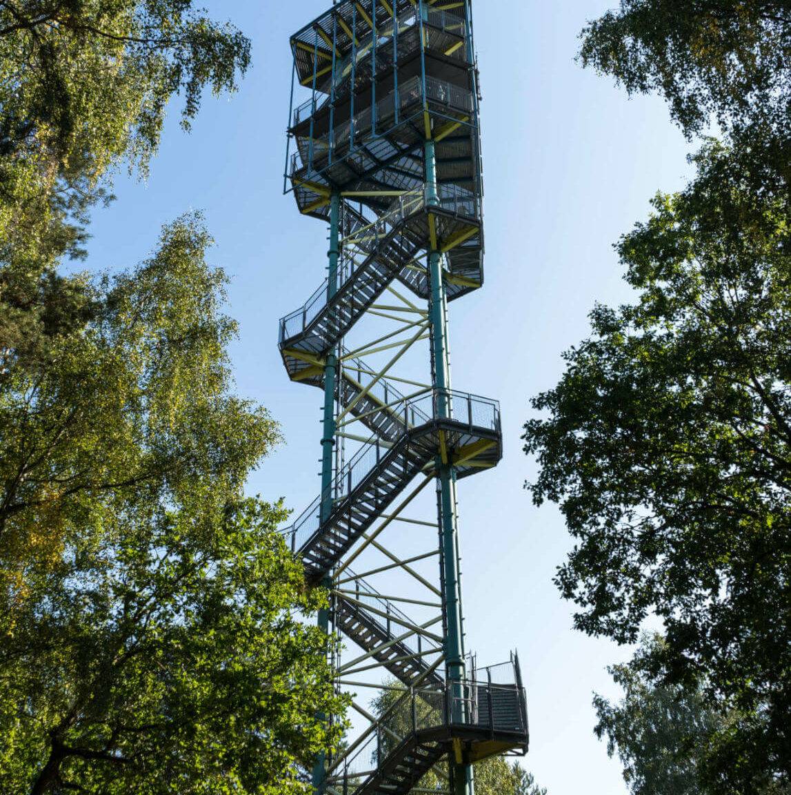 Aus der Froschperspektive erkennt man den Käflingsbergturm, ein mit einer Stahl-Wendeltreppe gebautes Konstrukt, das in Müritz als Aussichtsplattform dient, links und rechts vom Turm stehen große Bäume.