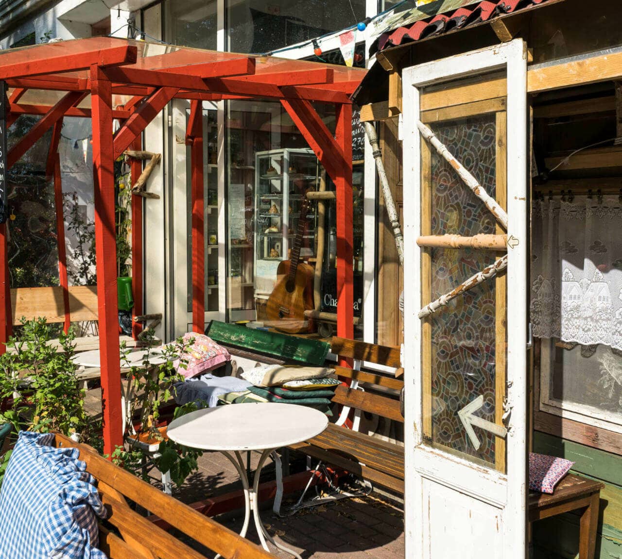 Den Eingang eines kleinen, alternativen Cafés in Rostock in der Kröpeliner-Tor-Vorstadt ziert eine rote Pergola, Holzbänke, Kissen und weiße Tische, eine alte weiße Holztür mit Pfeil zeigt ins Café hinein.