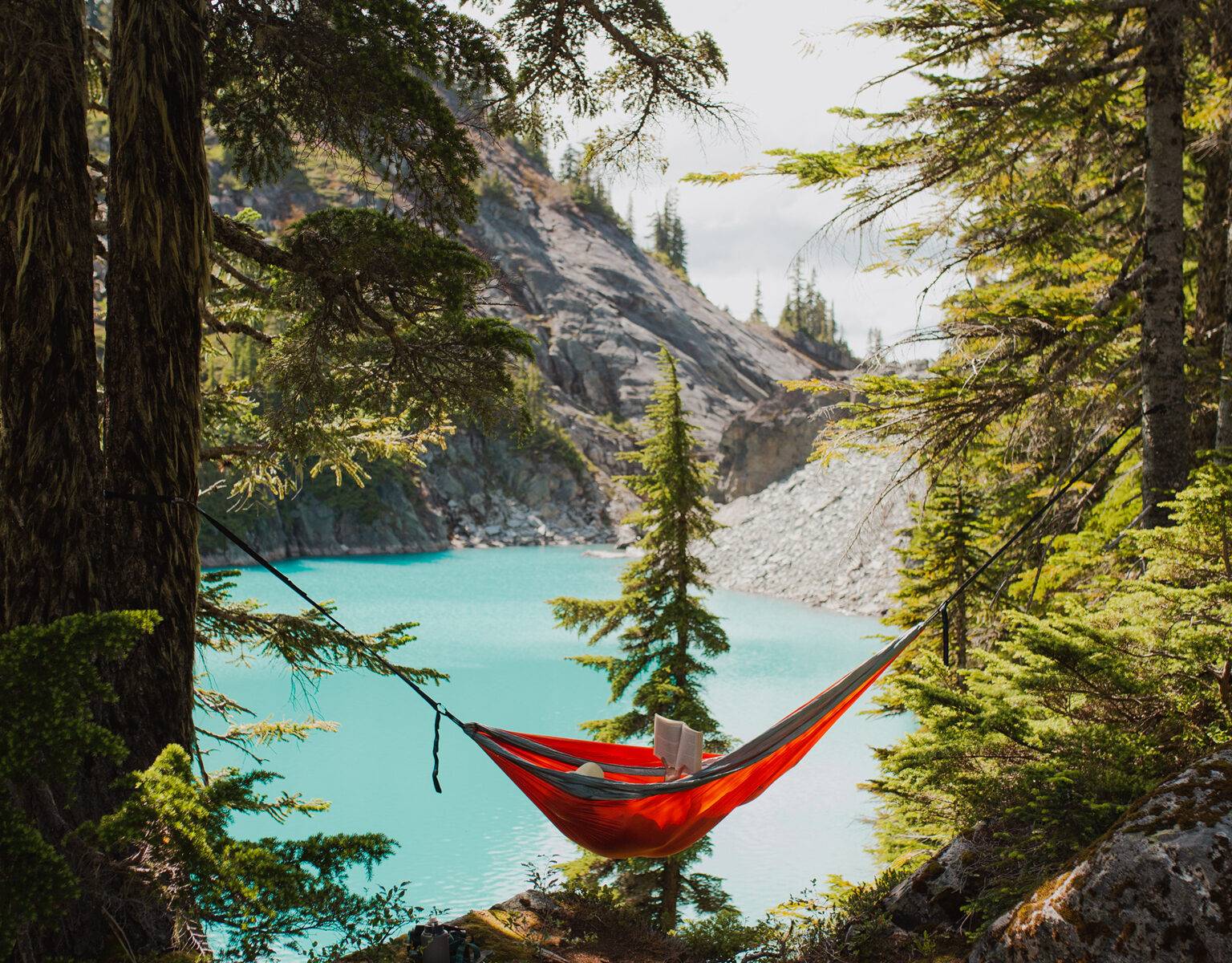 In einem Wald vor einem türkisblauen See liegend ist eine rote Hängematte zwischen zwei Baumstämmen weit aufgespannt, darin liegt jemand der Sonnenhut trägt und ein Buch liest.