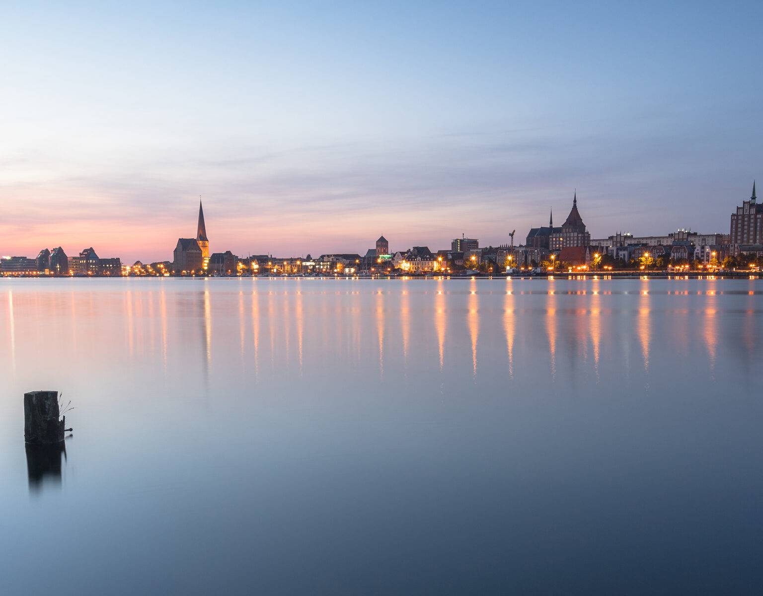 Die Skyline von Rostock liegt mit Lichtern erleuchtet vor dem Wasser im Sonnenuntergang, der Himmel und das Wasser sind pink gefärbt.