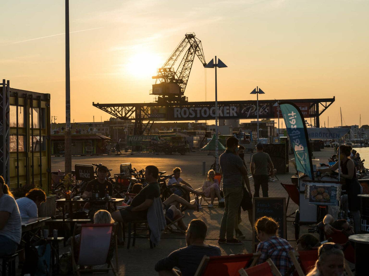 Im Sommer einen Besuch wert: Menschen sitzen in einer Bar am Hafen von Rostock bei Sonnenuntergang, im Hintergrund befindet sich ein Schiffskran.