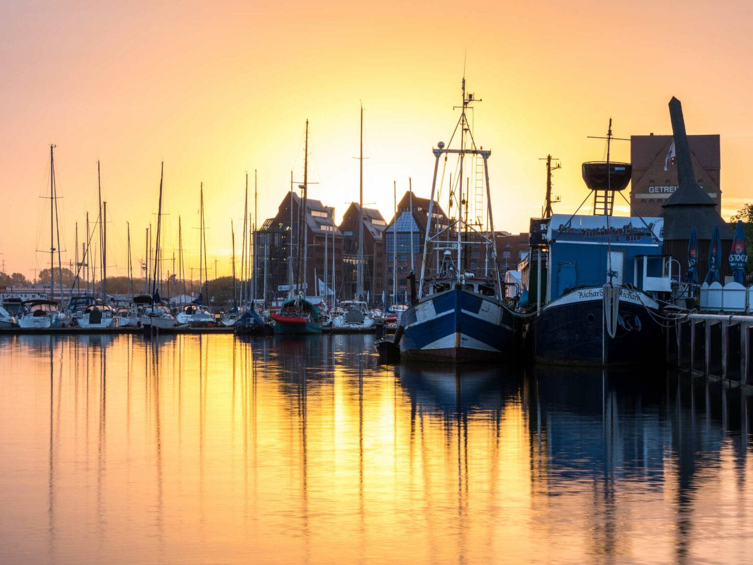 Diverse Schiffe, Yachten und Dampfer liegen bei Sonnenaufgang im Rostocker Stadthafen. die untergehende Sonne spiegelt sich im Wasser.