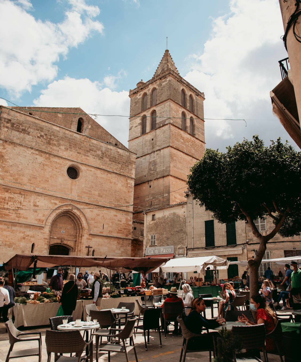 Ein gut besuchtes Café auf dem Wochenmarkt von Sineu auf Mallorca mit einem Gemüsestand und altertümlichen Gebäuden im Hintergrund.
