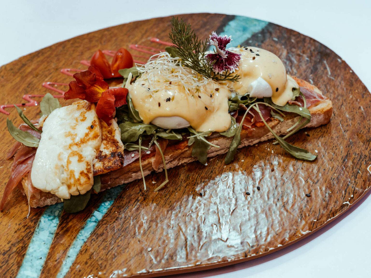 Beim Crustina Eggullera, was in der Eixampeling Brunch Café & Bar in Barcelona serviert wird, gibt es eine getoastete Scheibe Brot mit Serrano Schinken, pochierten Eiern, Sauce Hollandaise, Rucola und Tomaten.