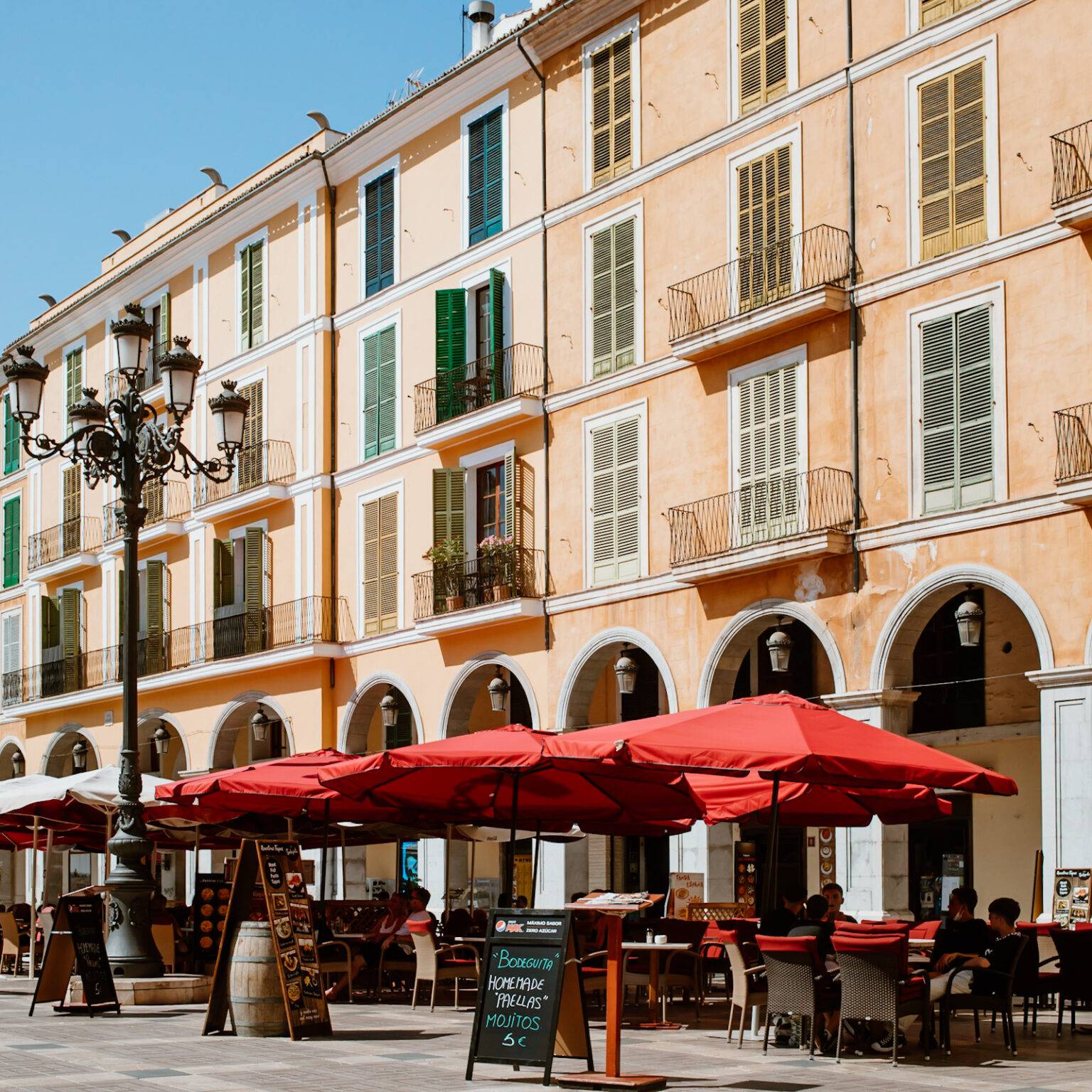 Der große Platz Plaza Mayor mit sandfarbigen Häuserwänden und roten Restaurantschirmen in Palma.