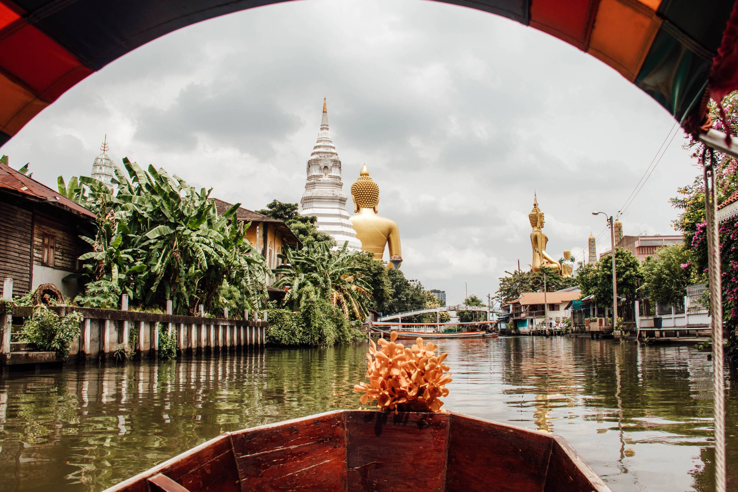 Blick von einem Boot auf einem breiten Fluss in Bangkok mit goldenen Skulpturen im Hintergrund.