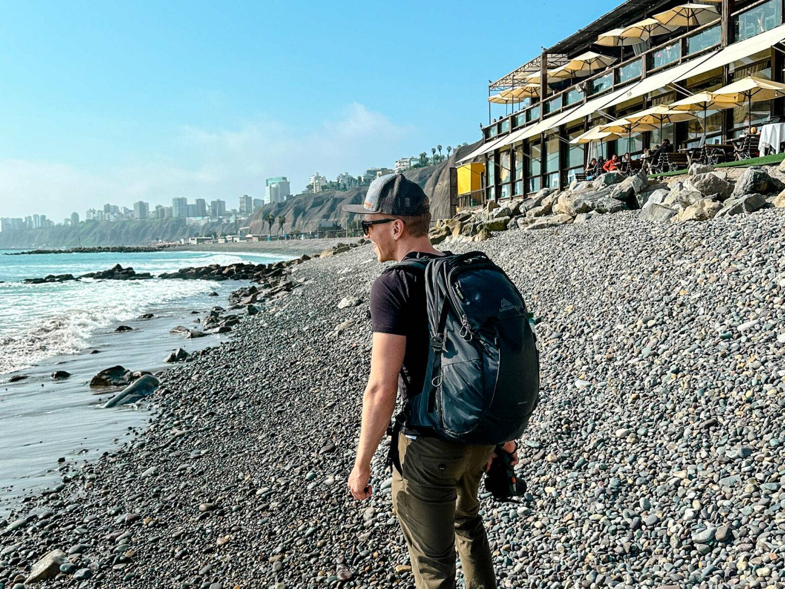 Reiseblogger Tom von traveloptimizer steht mit Rucksack an einem kieselsteinigen Strand in Lima.