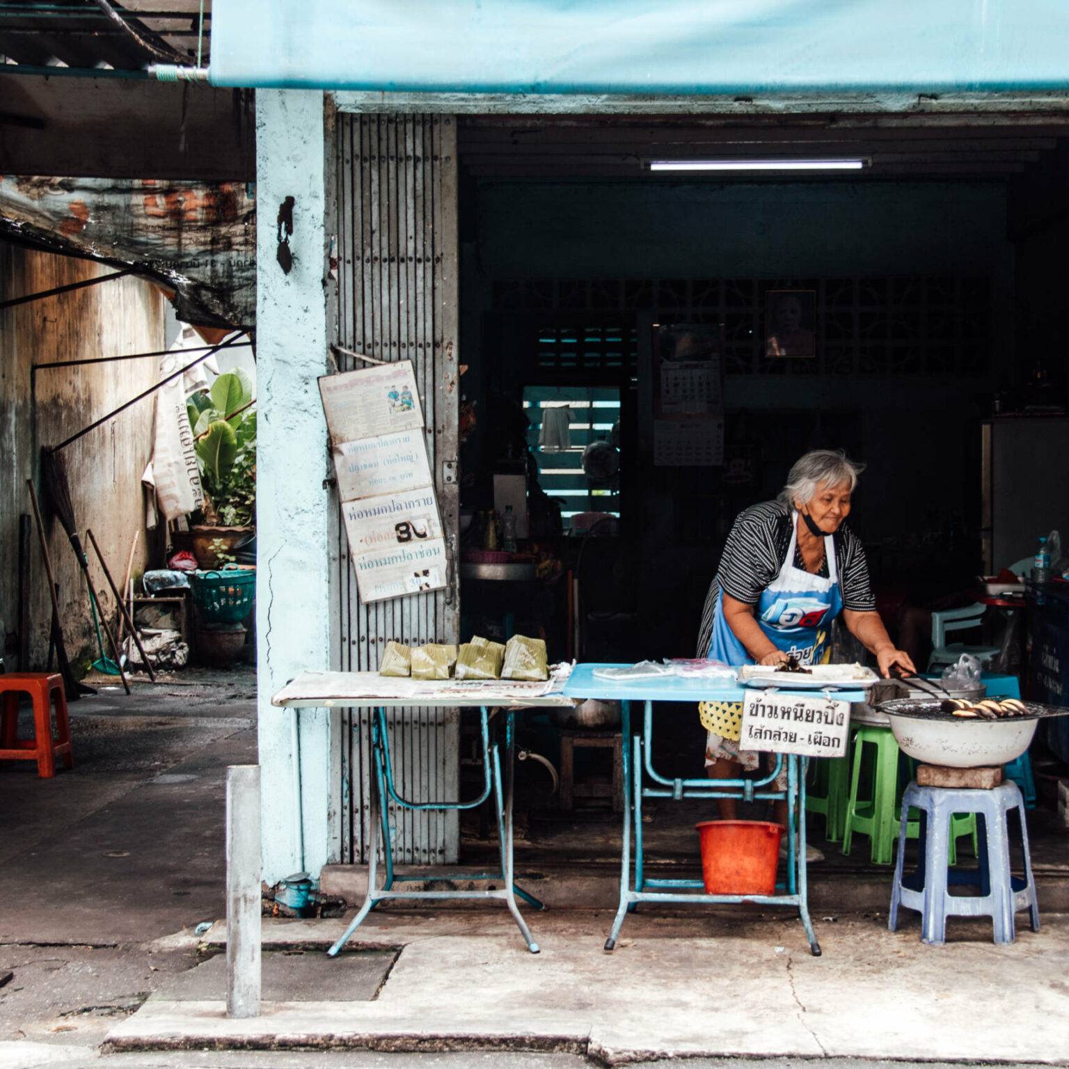 Eine ältere Dame steht hinter einer Streetfood Garküchen in Bangkok und kocht.