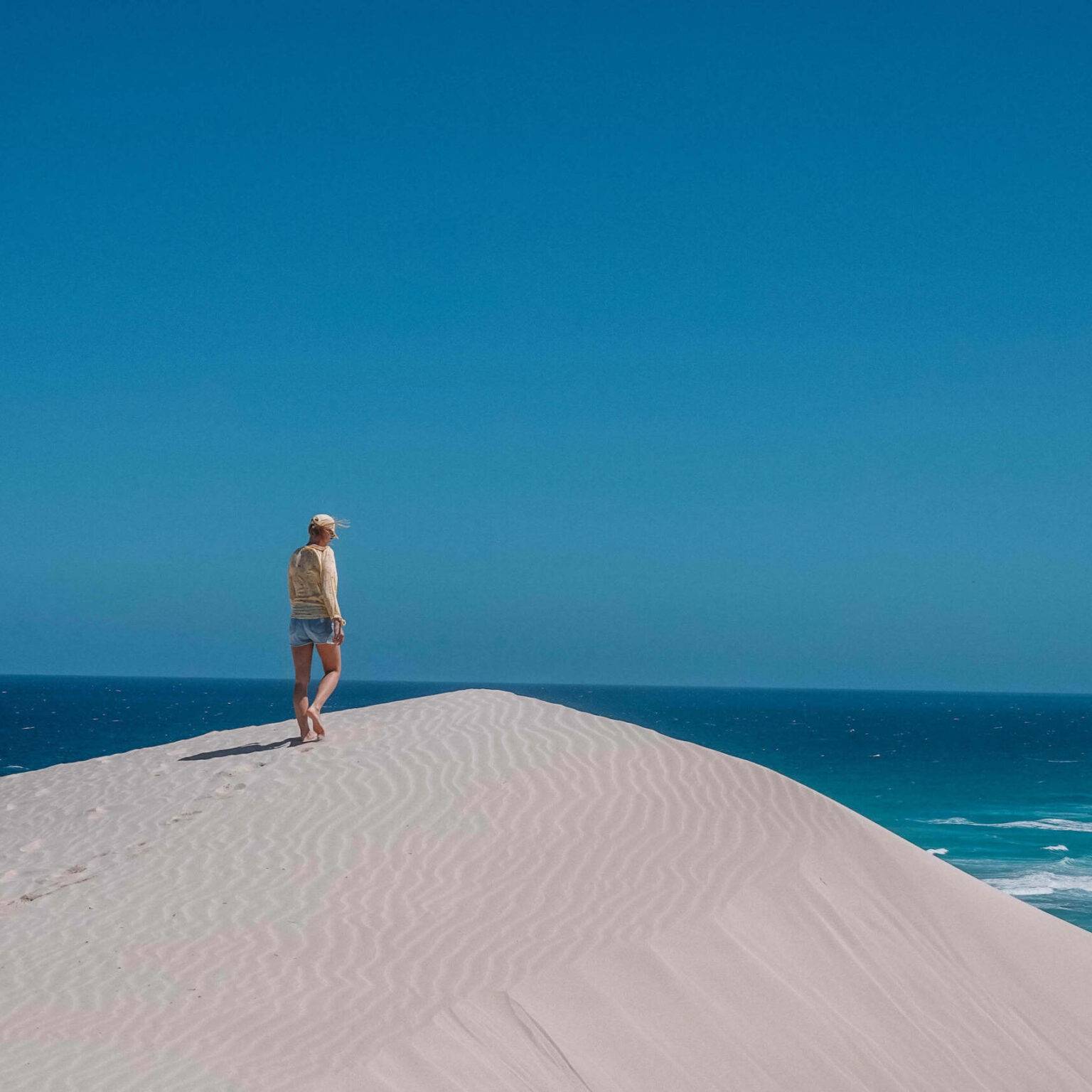 Bloggerin Nina steht auf einer Düne in weißem Sand mit türkisblauem Meer und einem blauen Himmel im Hintergrund.