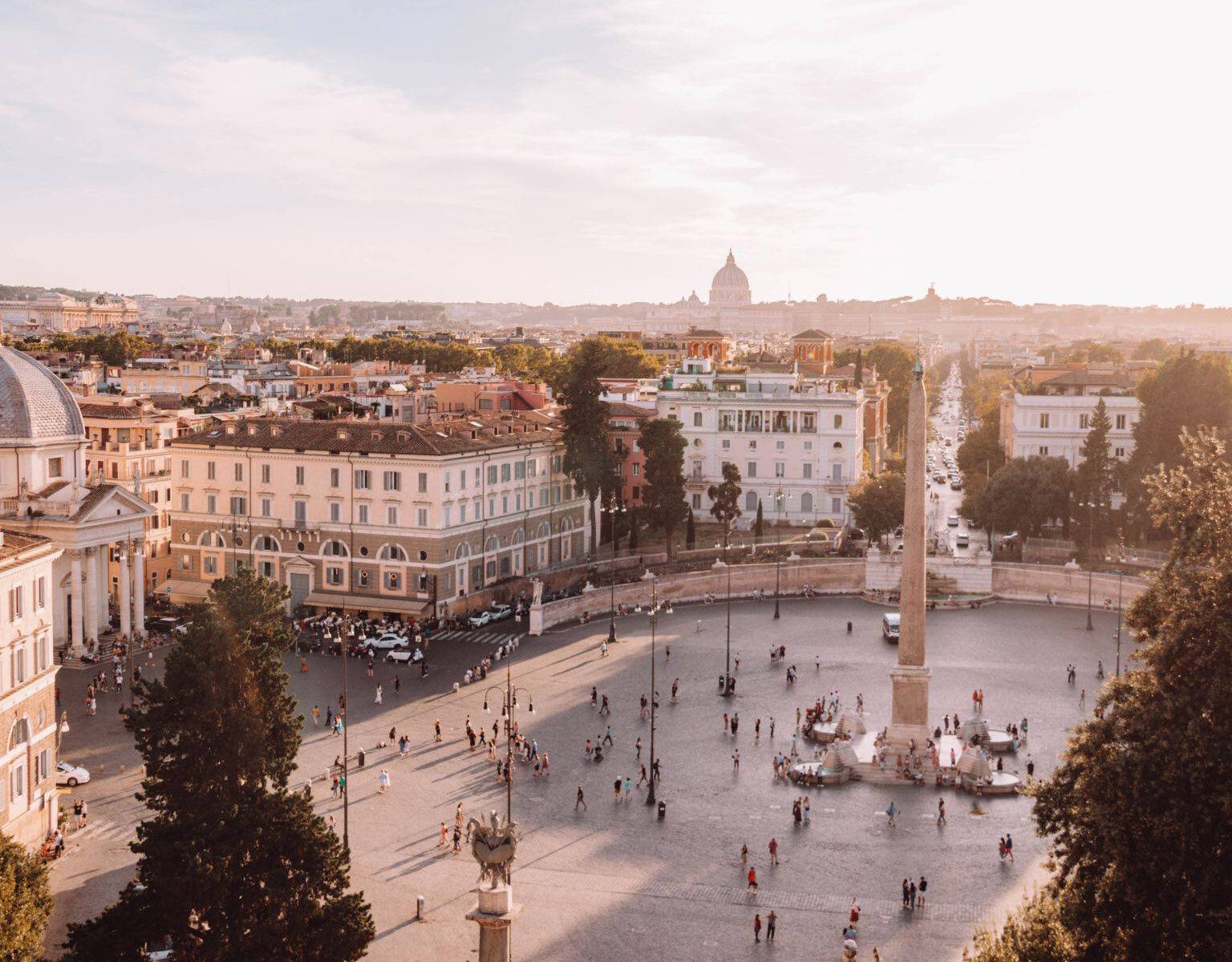 Eine Säule steht inmitten des Piazza del Popolo. Das Panorama im Hintergrund zeigt Rom in der Abendsonne.