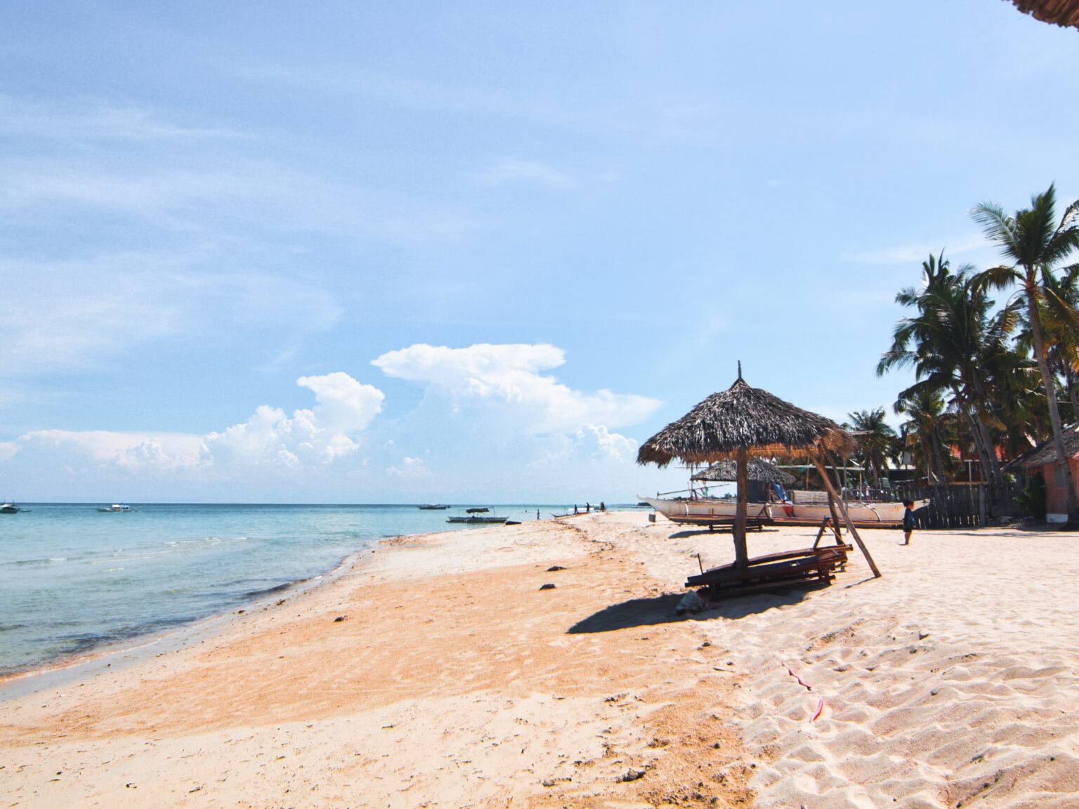 Ein Schirm aus Palmenblättern spendet Schatten am dem schönen Strand mit feinem Sand und kleinen Fischerbooten im Hintergrund.
