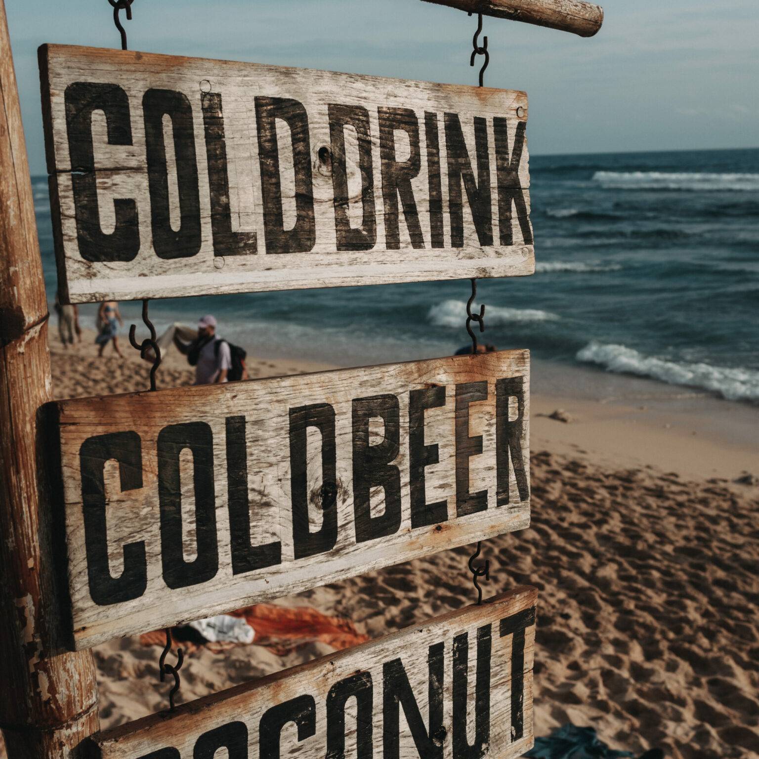 Ein Holzschild mit der Aufschrift "Cold Drink, Cold Beer, Coconut" an einem Strand im Süden von Bali.