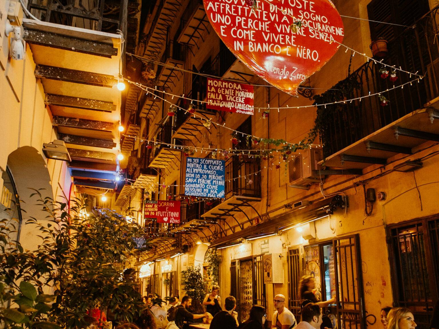 Eine Gasse in Palermo am Abend, Menschen sitzen draußen und essen.
