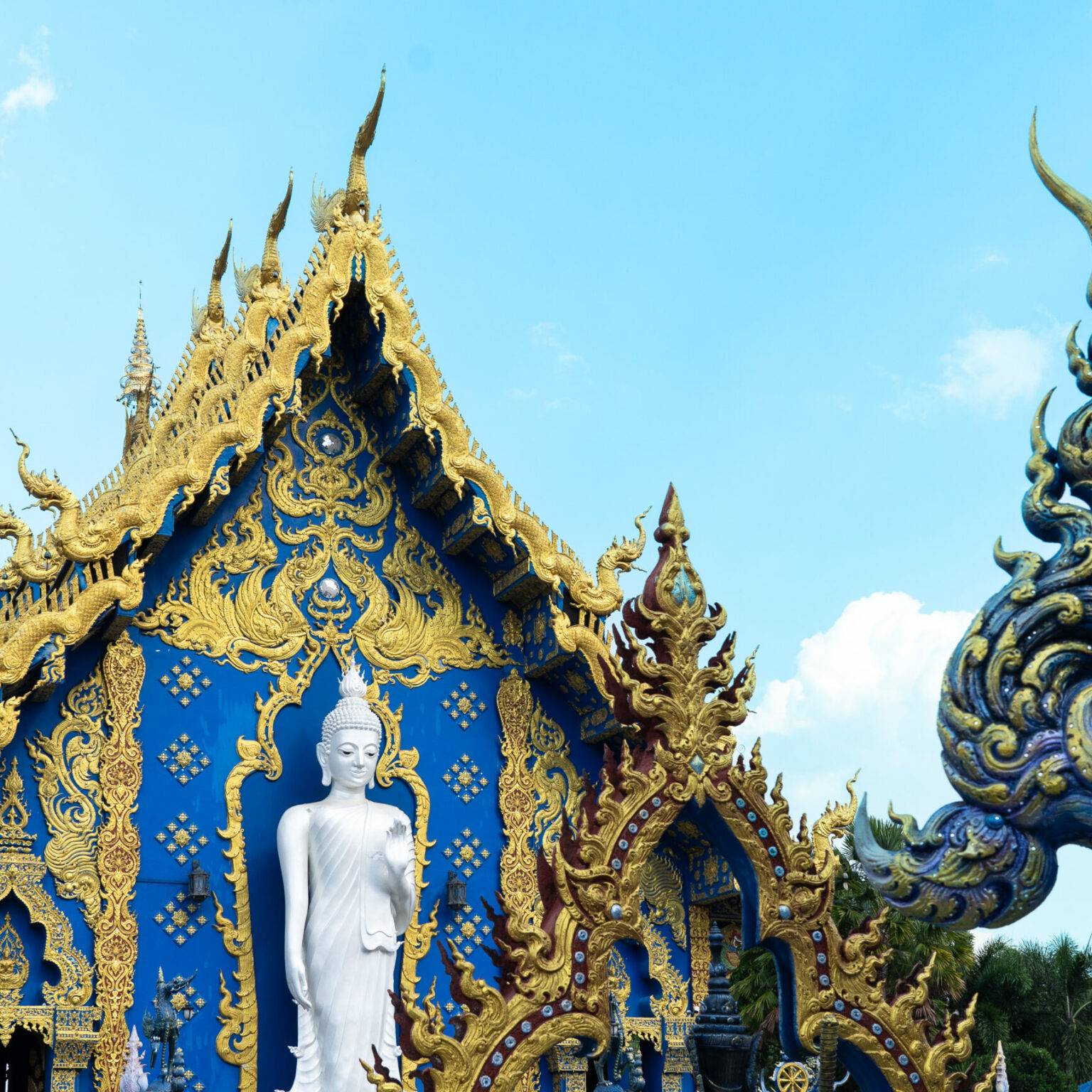 Eine kleine, weiße Buddha-Statue steht vor einer Wand des reich verzierten blauen Tempel.