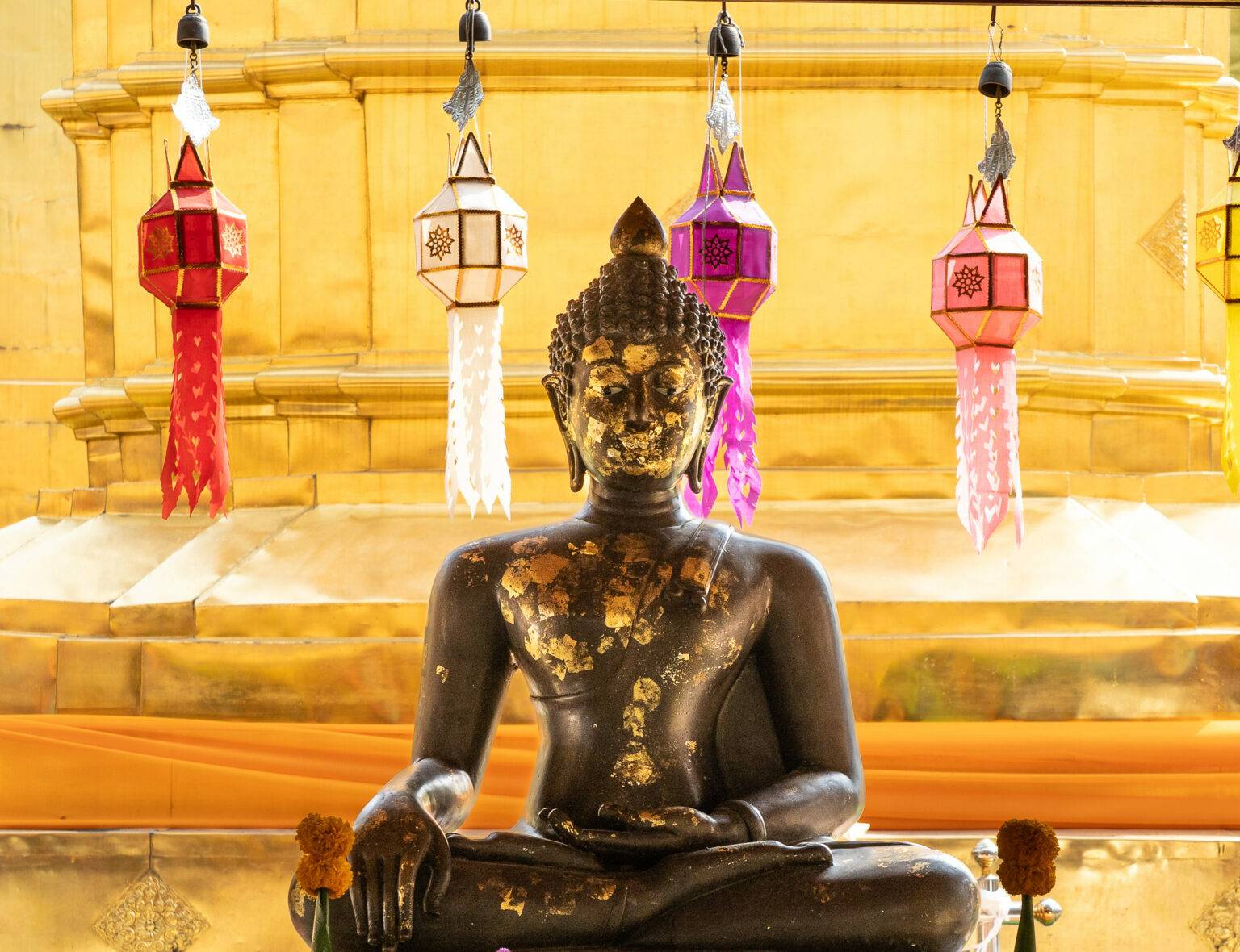 Eine Buddha-Statue sitzt zwischen Holzpfosten des Wat Pra Singh Tempels, an denen bunte Girlanden herabhängen.