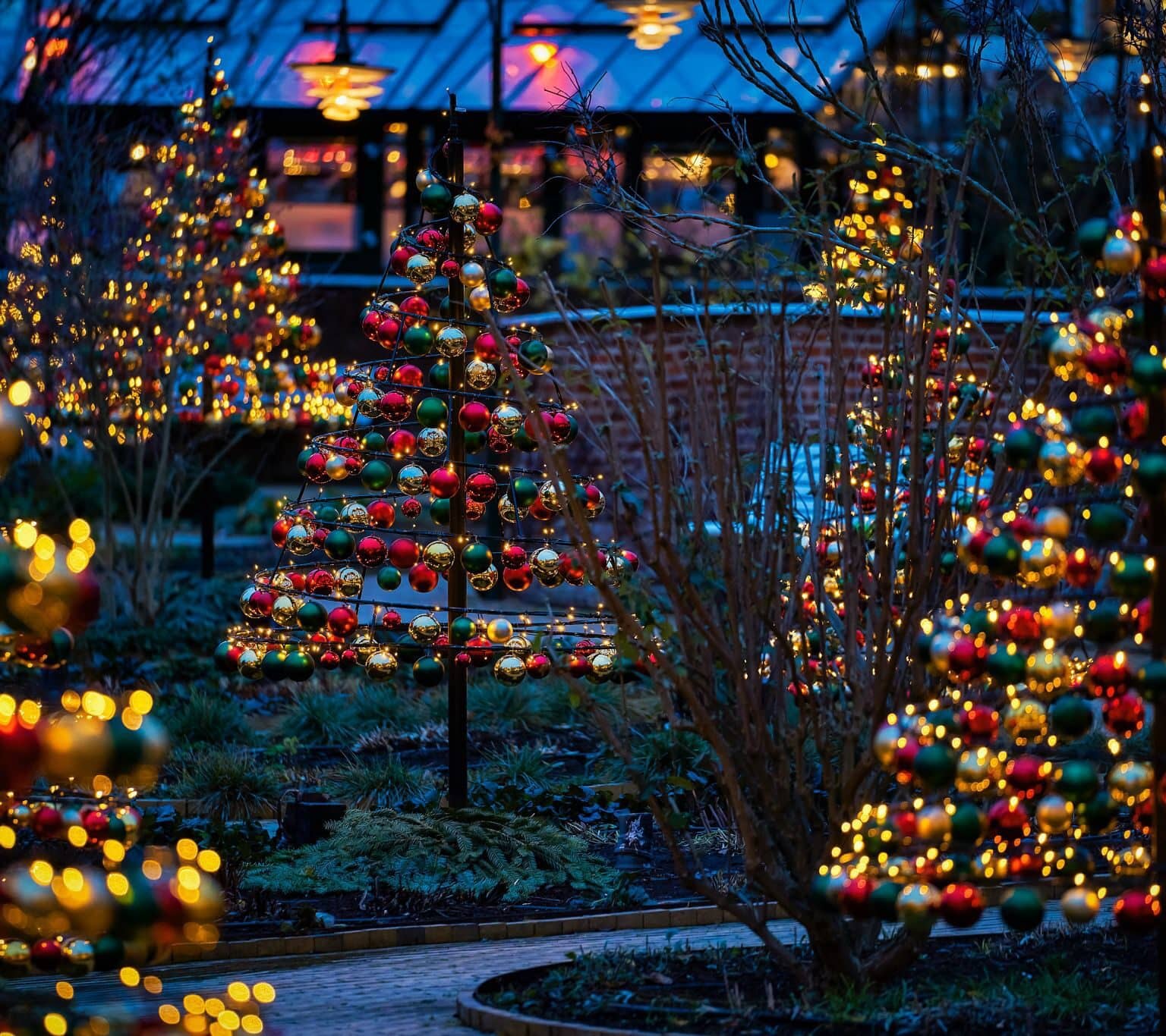 Mehrere kleine Weihnachtsbäume aus Weihnachtskugeln und Lichterketten säumen einen schmalen Pfad im Tivoli Freizeitpark.