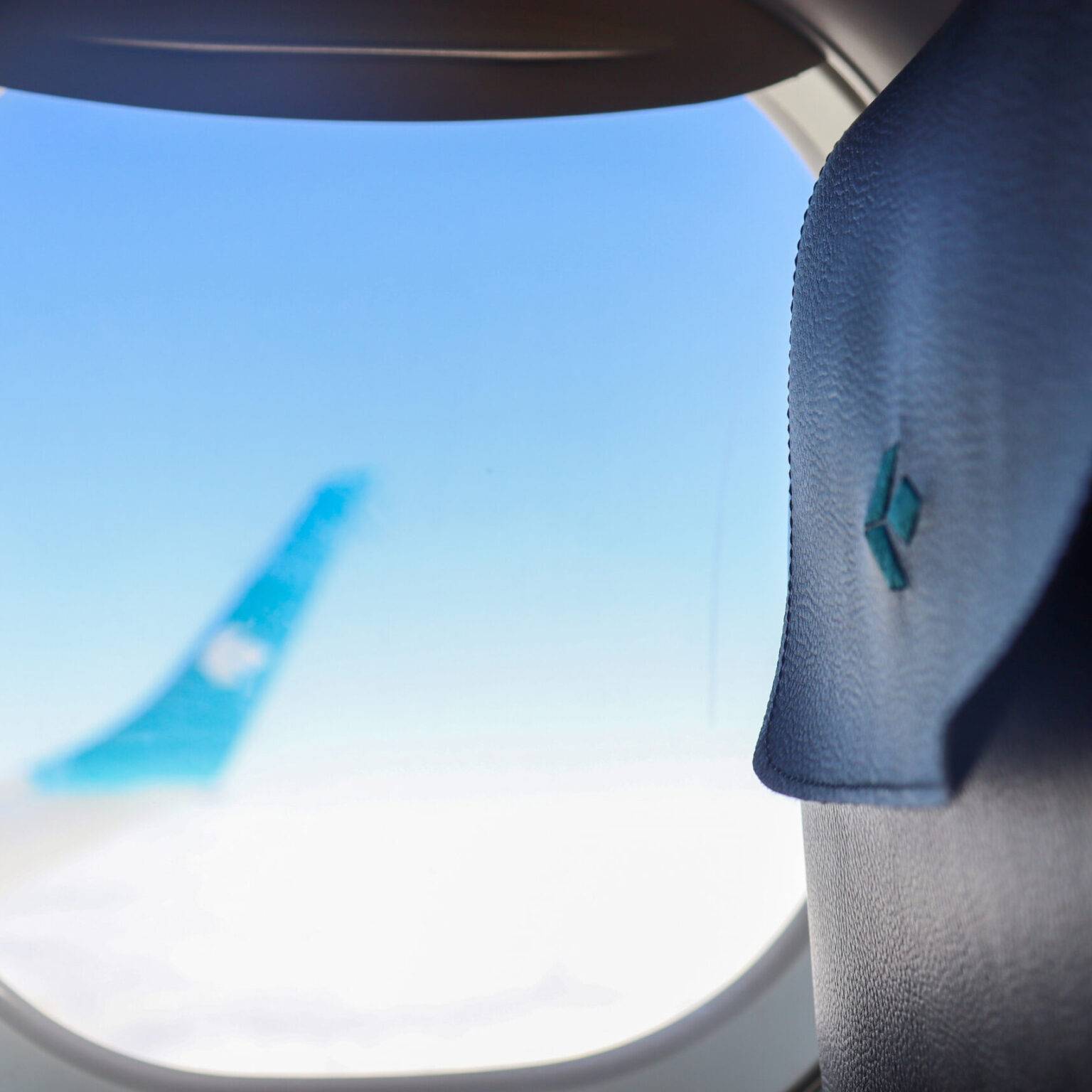 Das Kopfteil eines Sitzes in einem Flugzeug von Air Dolomiti neben dem Flugzeugfenster. Verschwommen im Hintergrund die Tragfläche über den Wolken.