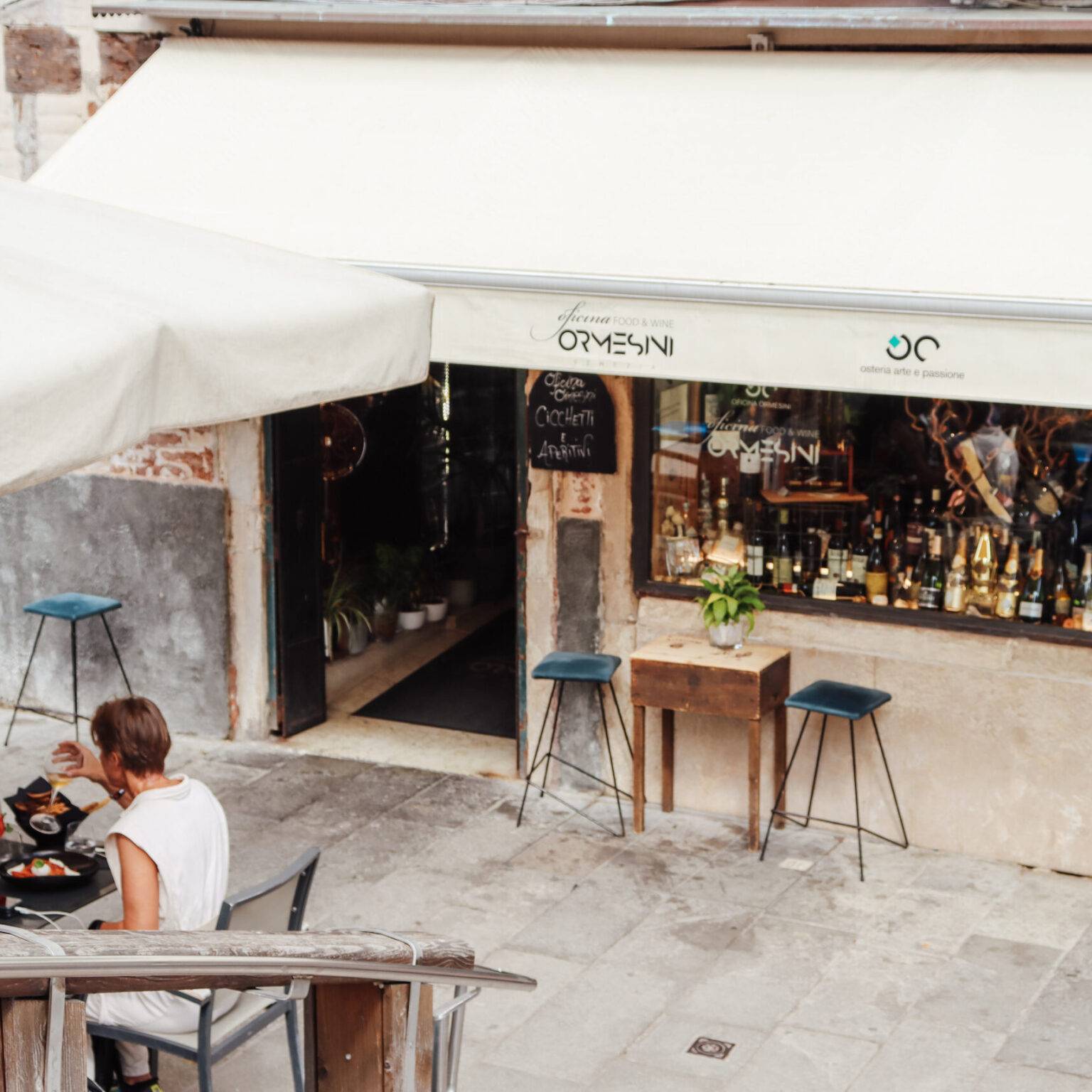 Die Fassade vom Restaurant Ormesini mit weißer Markise. Auf der Straße vor dem Restaurant sitzt eine Familie am Tisch und isst.