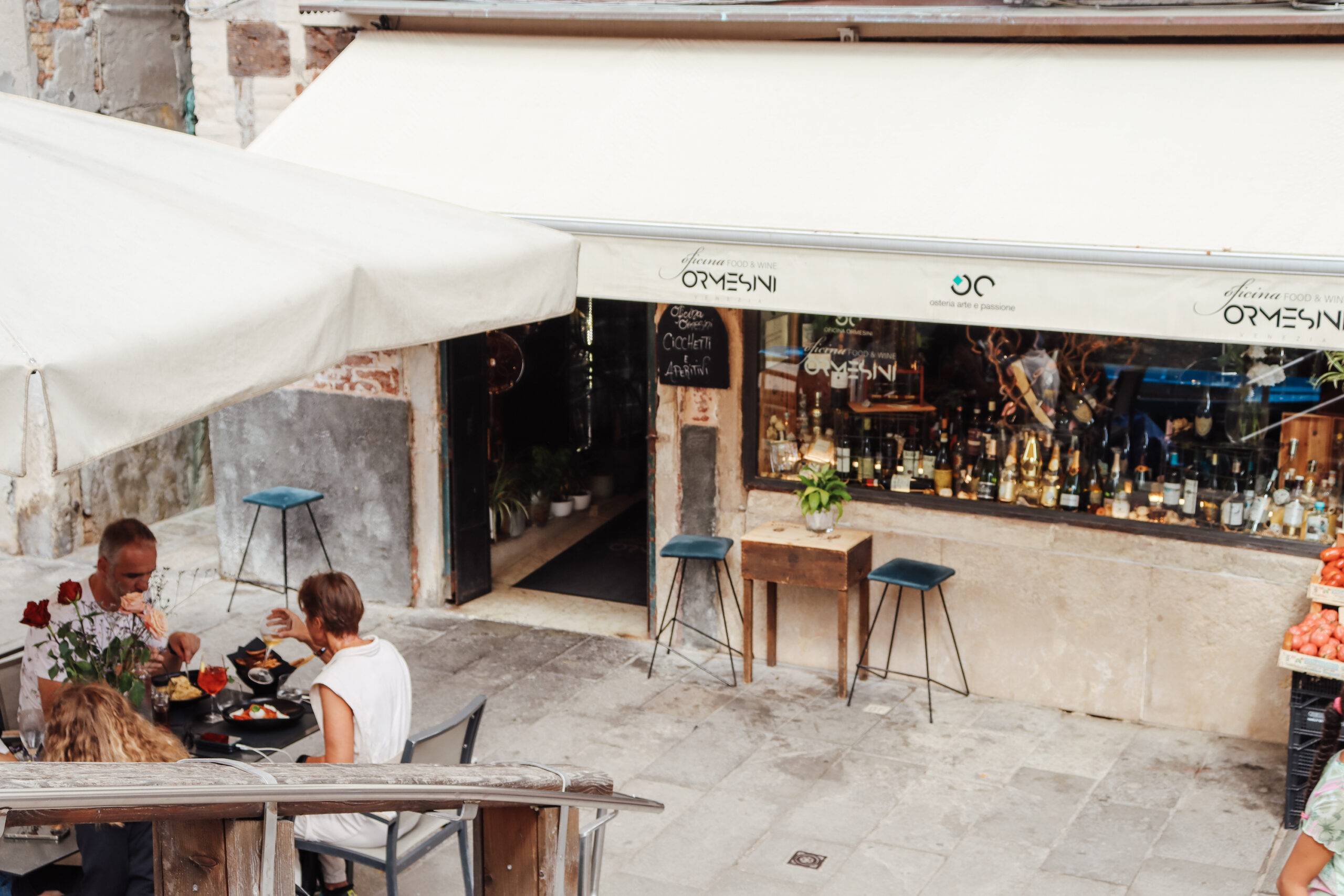 Die Fassade vom Restaurant Ormesini mit weißer Markise. Auf der Straße vor dem Restaurant sitzt eine Familie am Tisch und isst.
