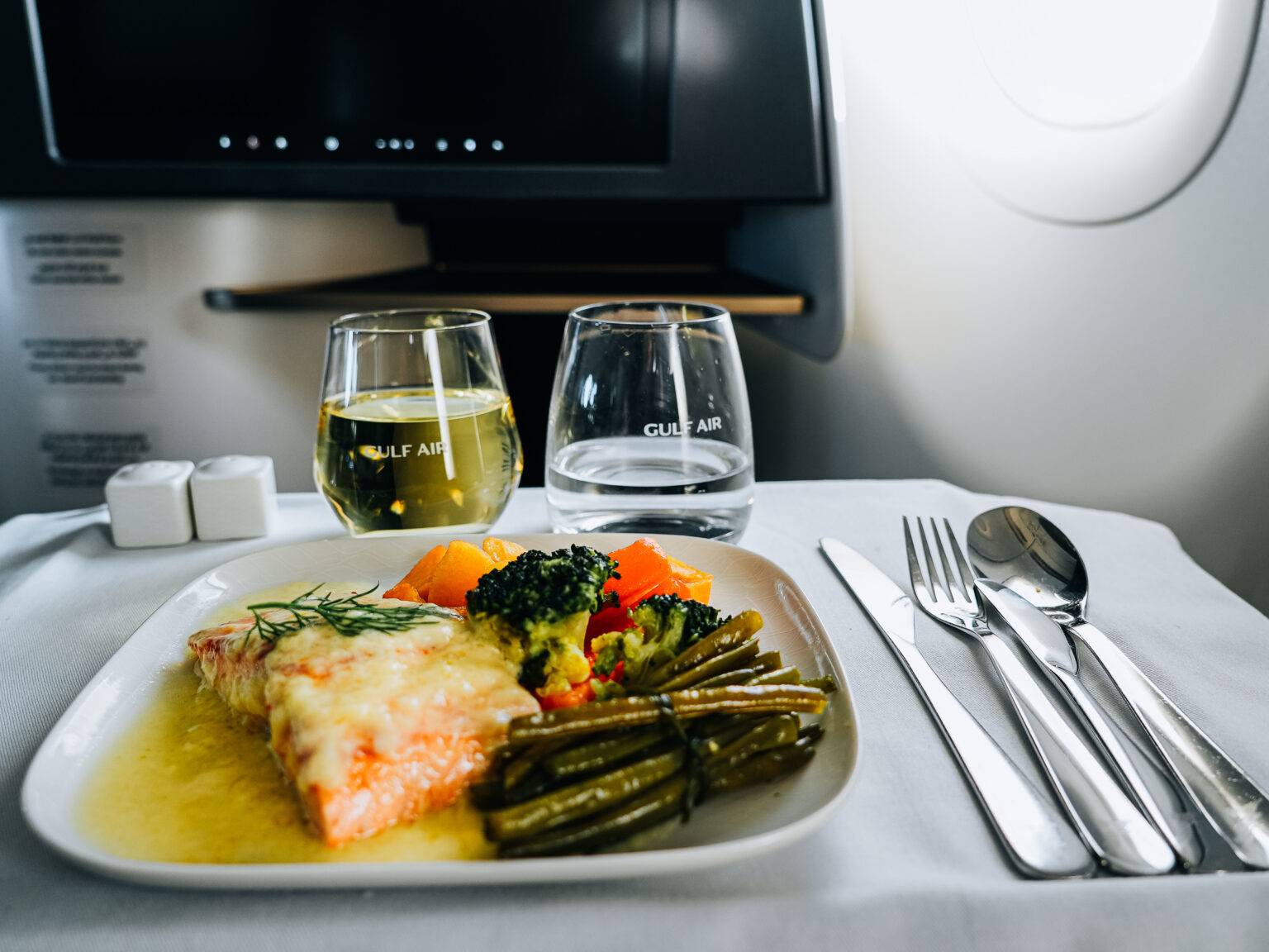 Ein Teller mit Fisch, Gemüse und Beilagen, begleitet von zwei Gläsern Wasser und Wein, auf einem Flugzeugtisch.