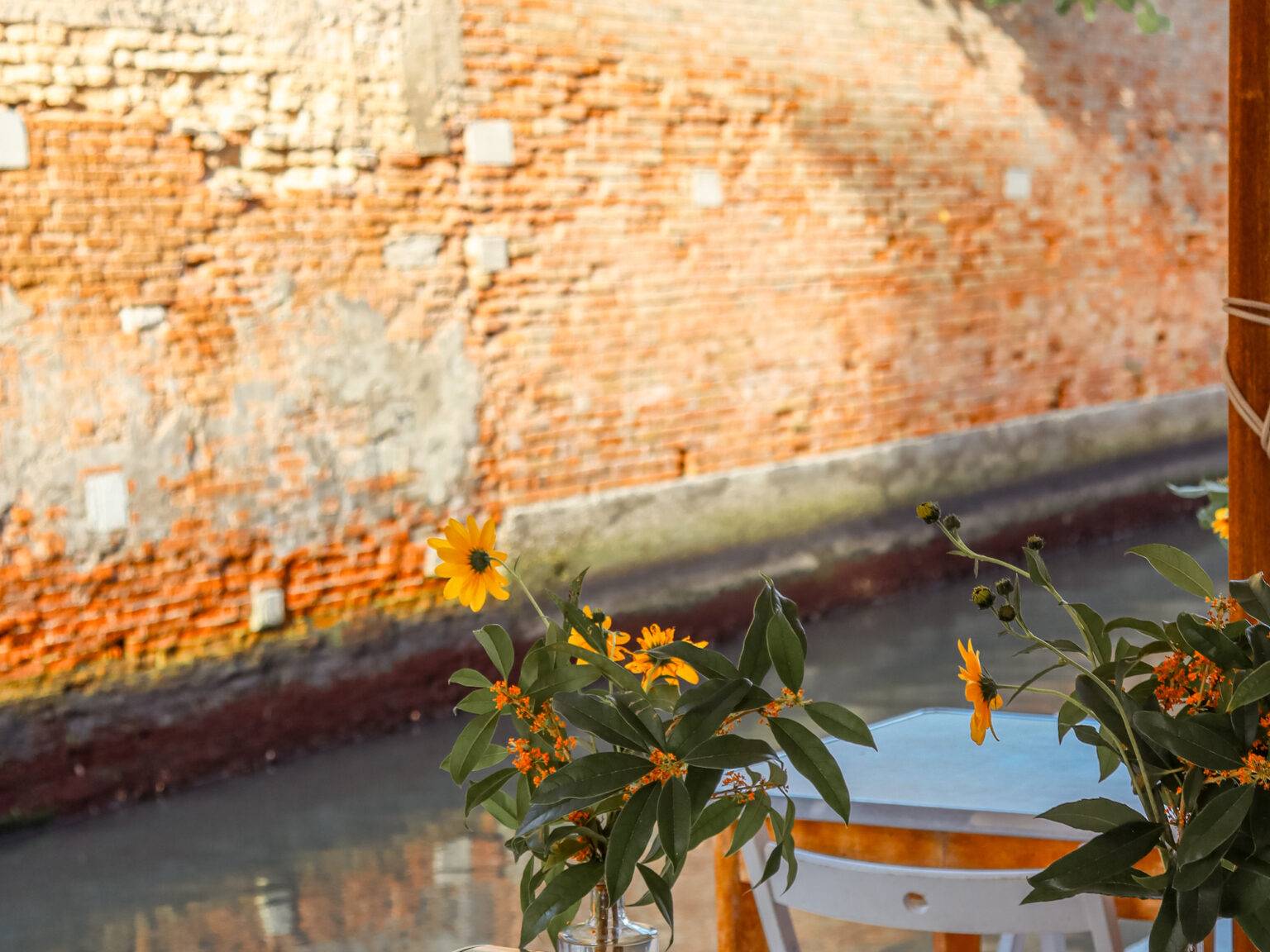 Unter einem Sonnenschirm stehen Tische und stühle direkt am Kanal. Auf den Tischen stehen Vasen mit frischen Blumen.