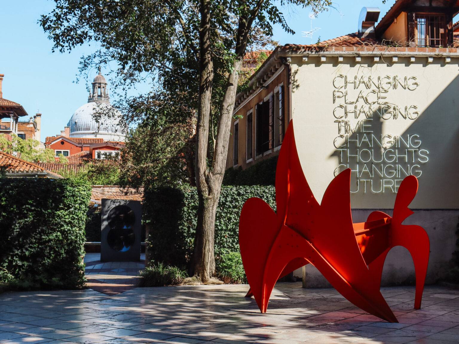 Der Garten der Peggy Guggenheim Collection mit einem roten Kunstwerk, gepflastertem Weg und grünen Hecken.