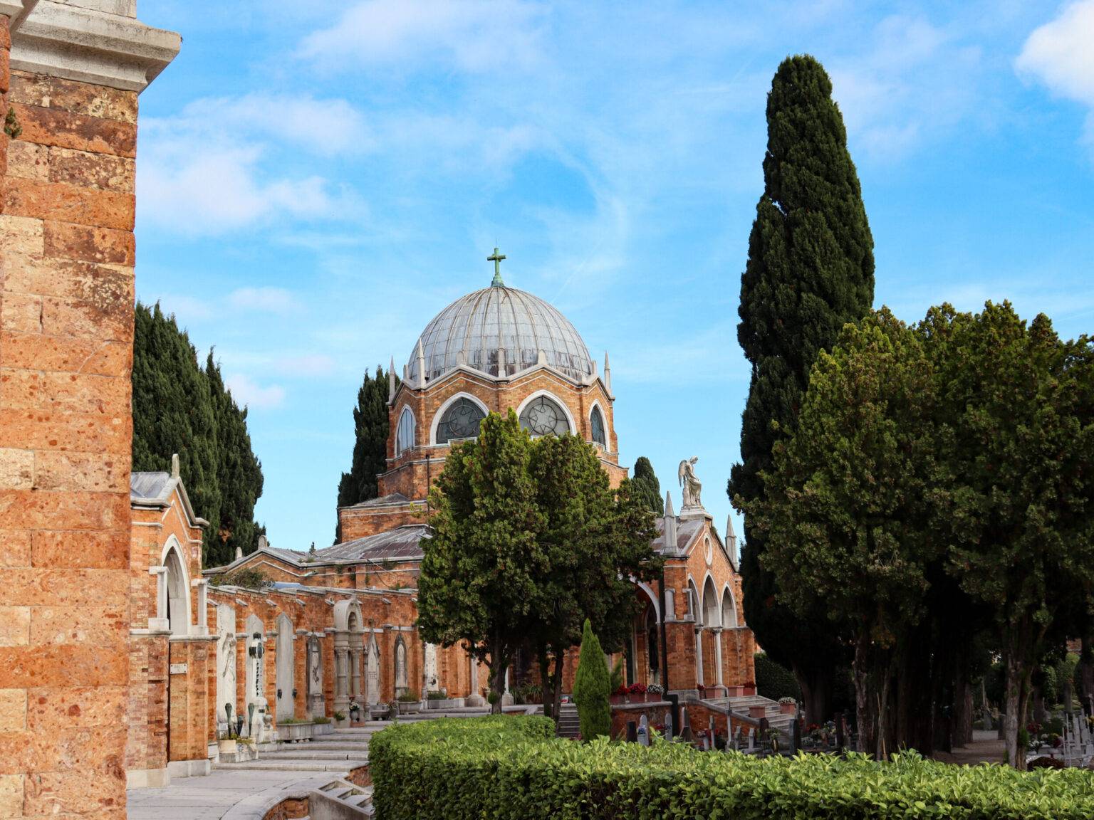 Die Fassade von San Michele mit Kirche, Grabsteinen, einer grünen Hecke und Bäumen.
