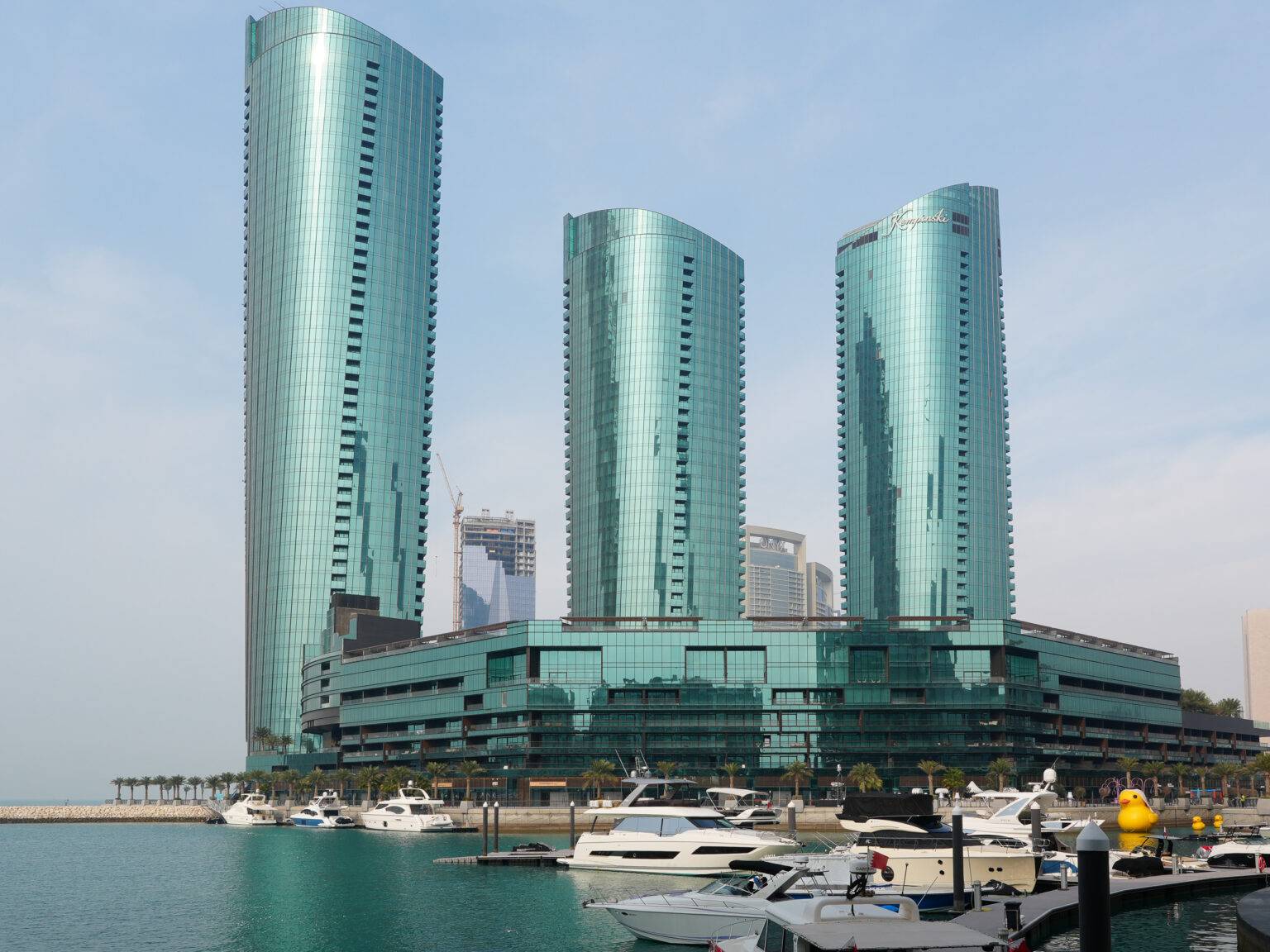 Drei moderne Glashochhäuser des Bahrain Financial Harbour, davor ein Yachthafen mit Booten.