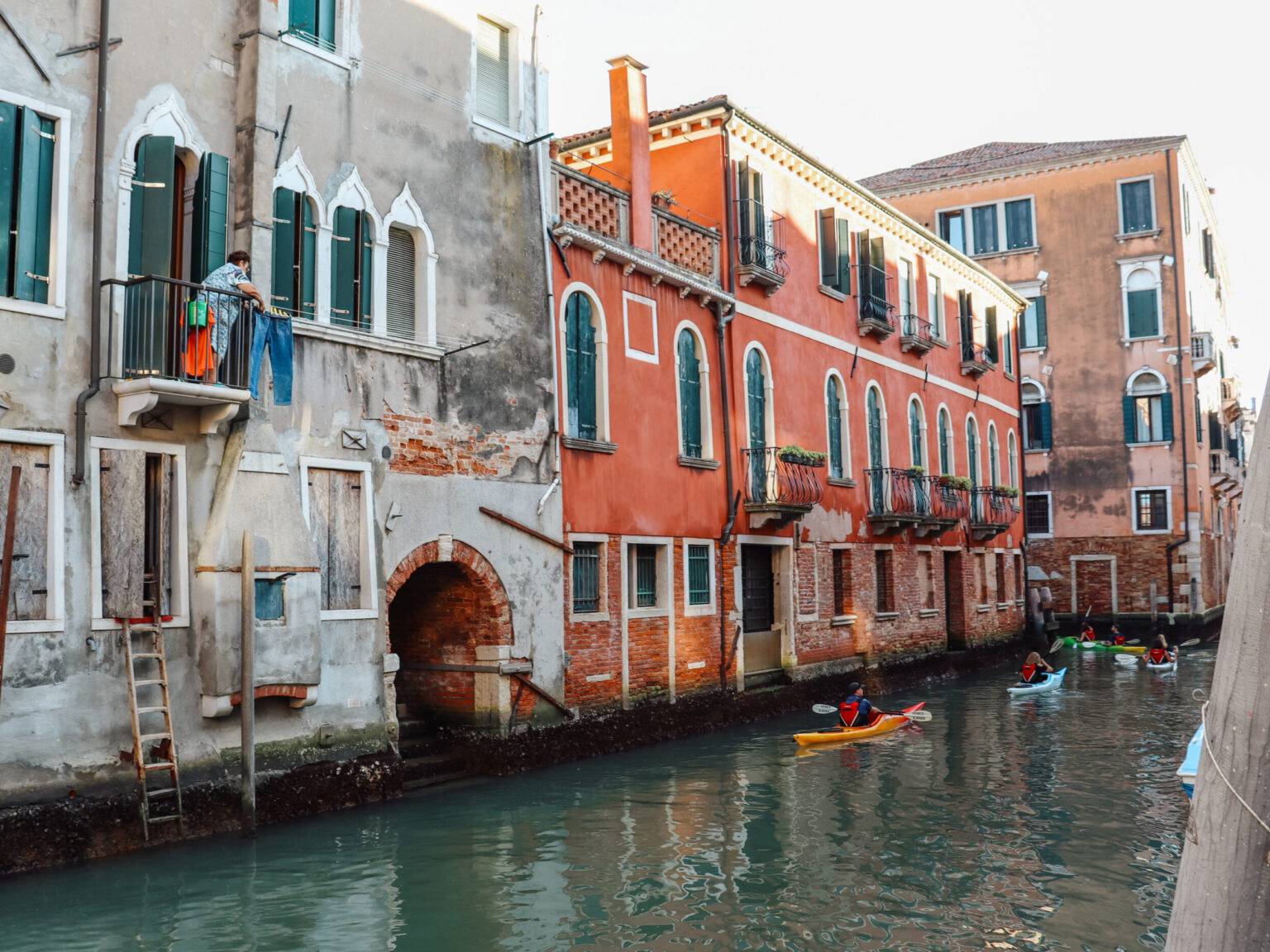 Auf einem Kanal in Cannaregio fahren Menschen in Kajaks. Auf einem Balkon an einem der Häuser steht eine Frau.