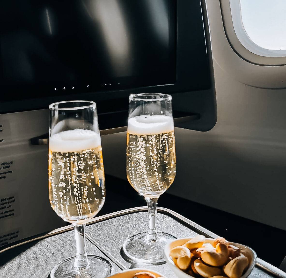 Zwei Gläser mit Sekt und zwei Schälchen mit Nüssen und Snacks auf einem Flugzeugtisch.
