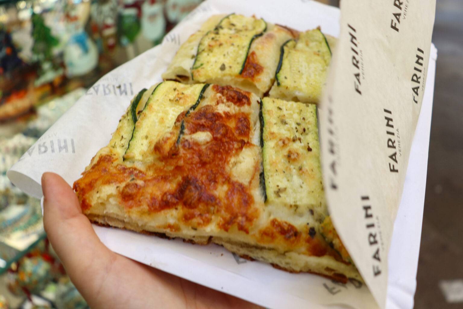 Ein Stück Pizza mit Zucchinistreifen auf die Hand.