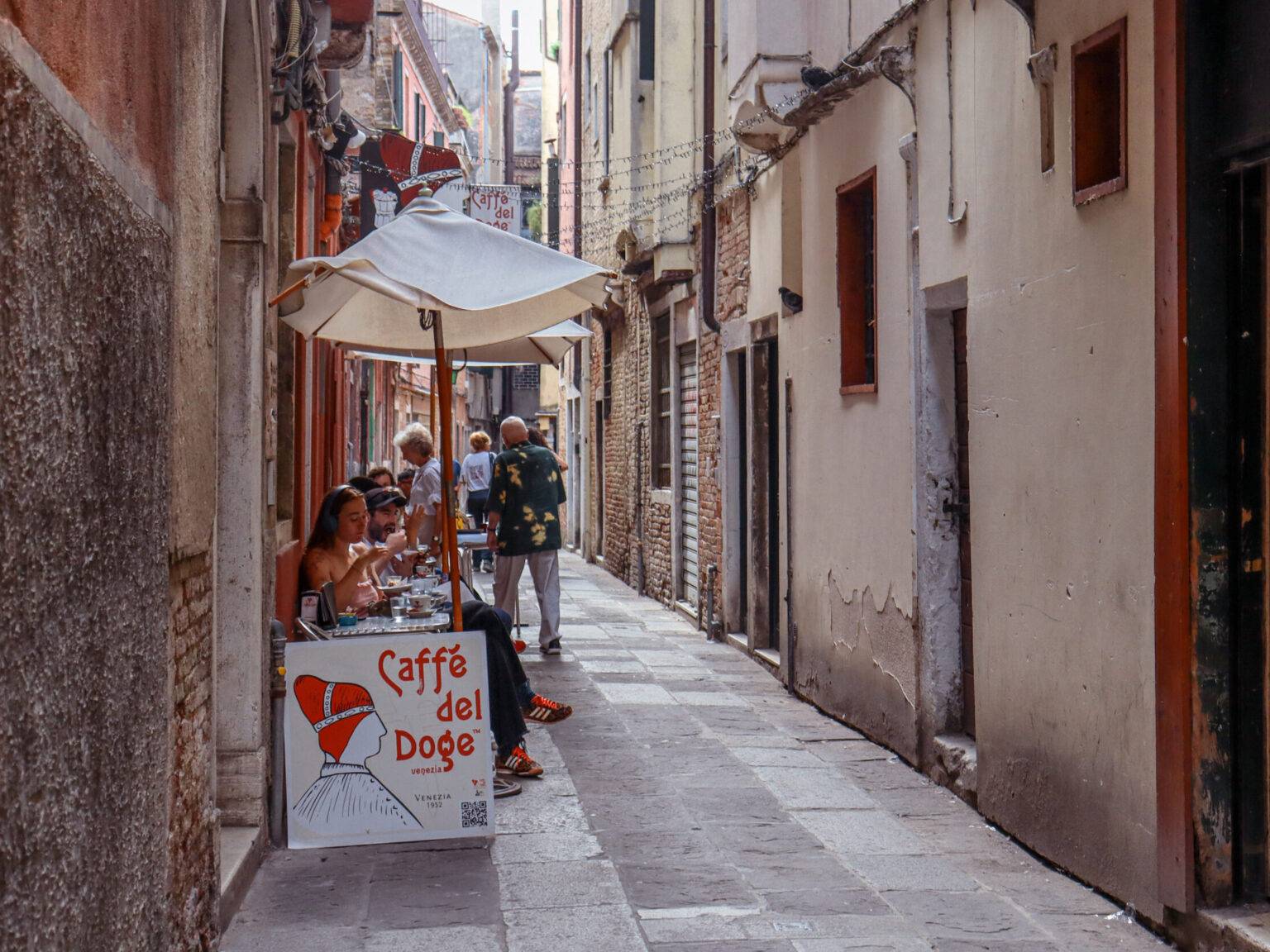 Eine schmale Gasse mit den außen stehenden kleine Tischen, Stühlen und Sonnenschirmen des Caffè del Doge in San Polo.