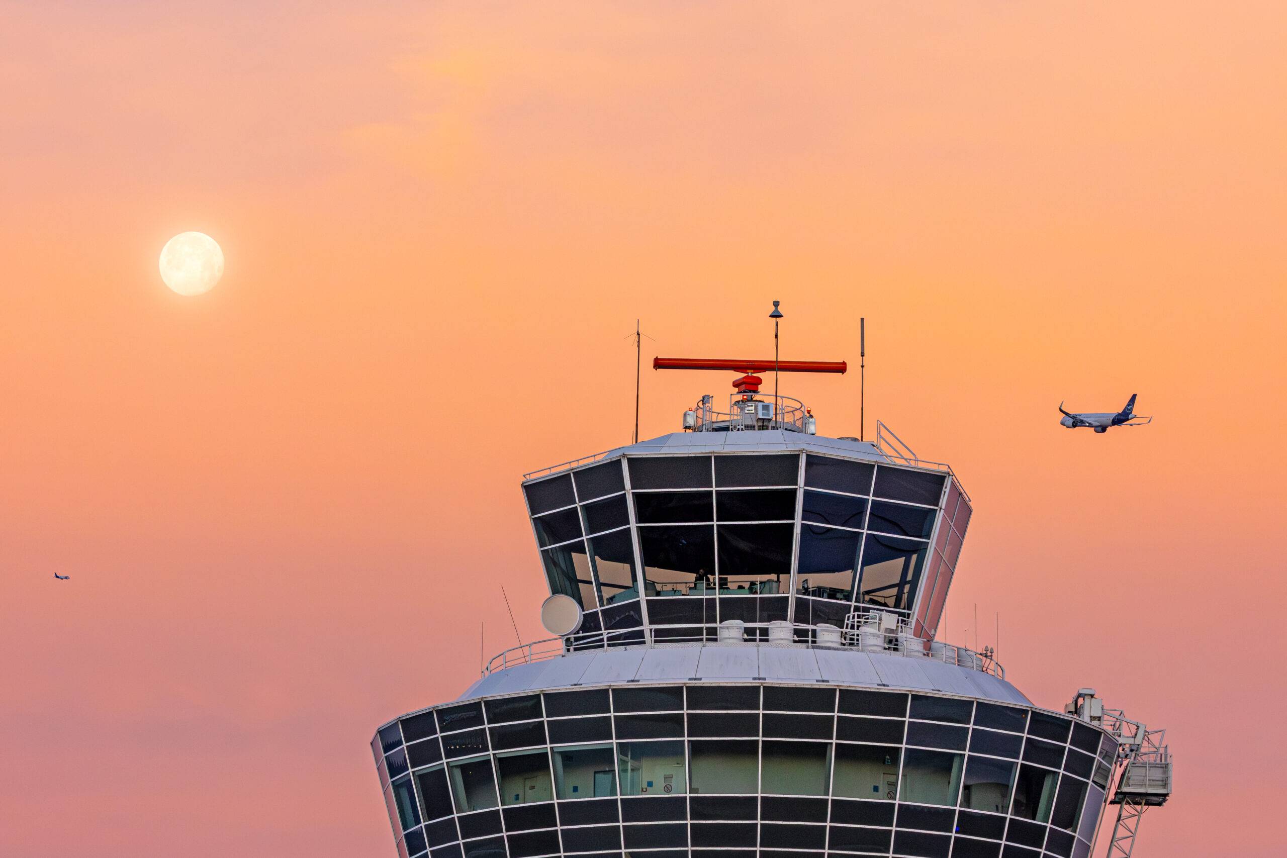 Der Tower des Flughafens München vor einem rot gefärbten Himmel, in den ein startendes Flugzeug aufsteigt.