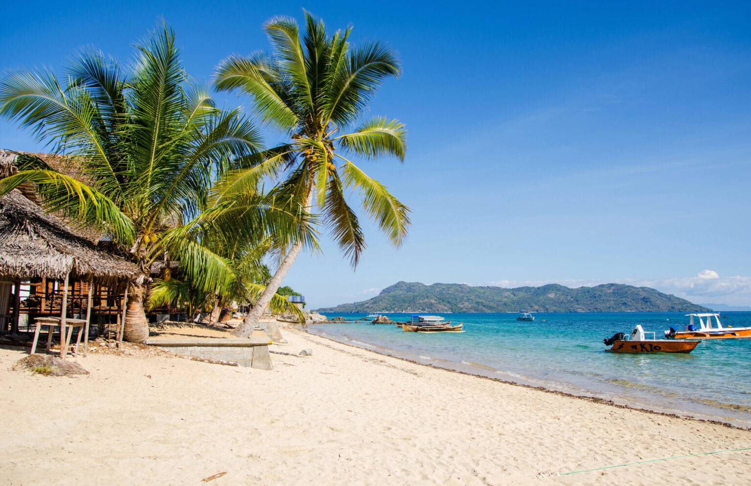 Kleine Ausflugsboote liegen im Meer vor dem palmengesäumten Strand der Insel Nosy Komba vor Madagaskar.