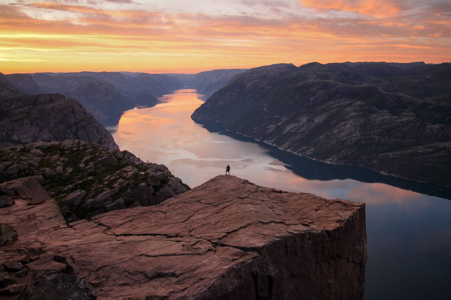 Ein Mann steht bei Sonnenaufgang auf dem Felsplateau Preikestolen. Zwischen den Felsen links und rechts fließt ein breiter Fluss.