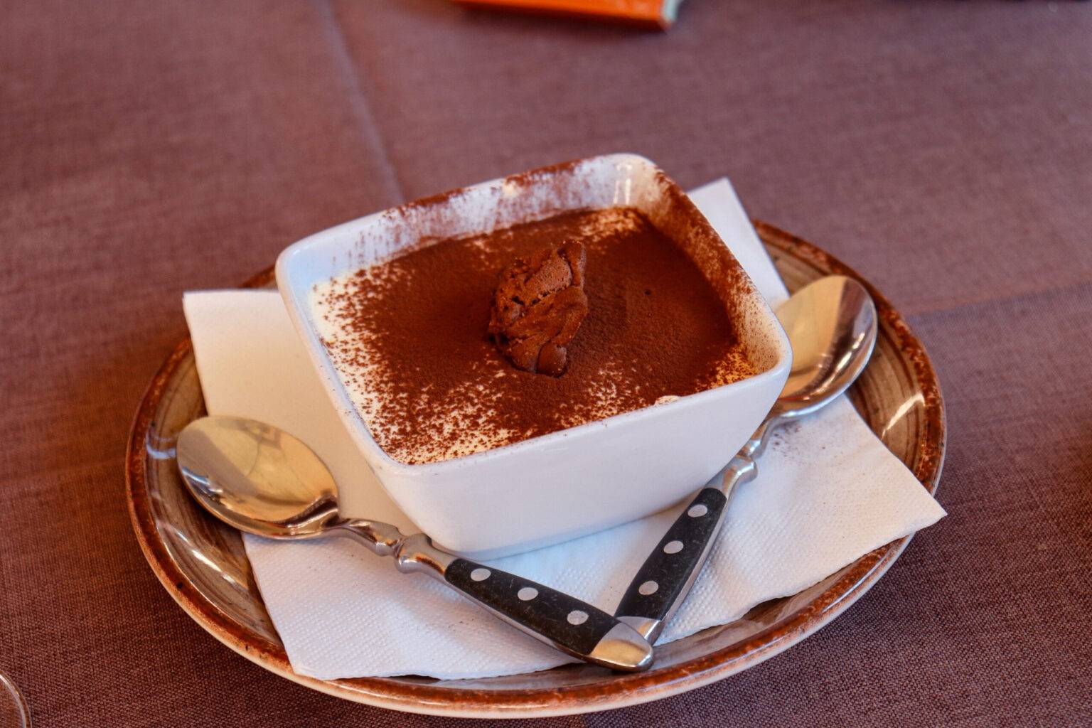 Auf einem Teller liegen zwei Löffel und ein Schälchen mit Kakao überstäubtem Tiramisu.