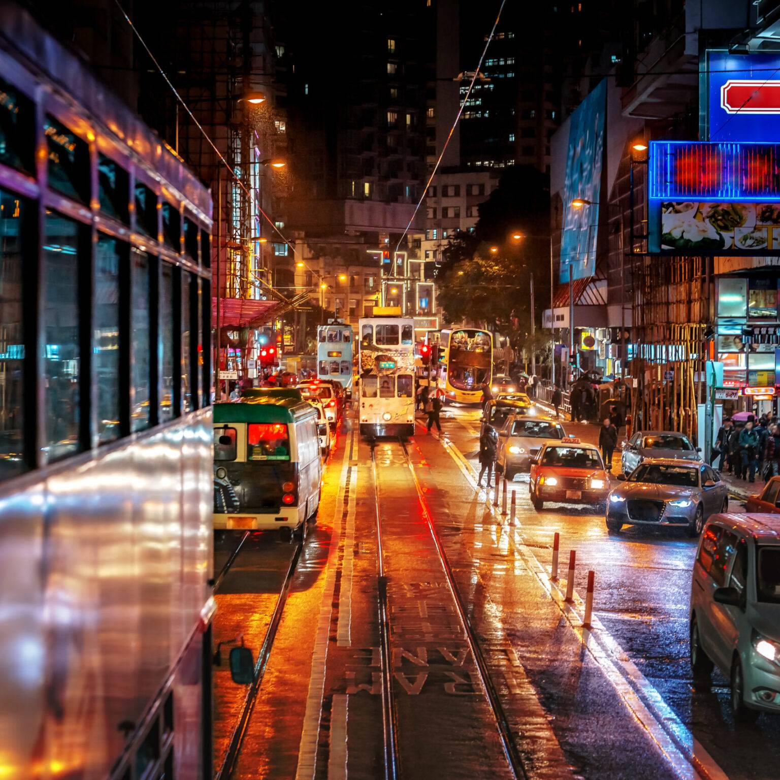 Eine mit Autos und Straßenbahnen stark befahrene Straße in Hongkong bei Nacht. An den Häuserfassaden links und rechts sind bunt leuchtende Raklame-Schilder angebracht.