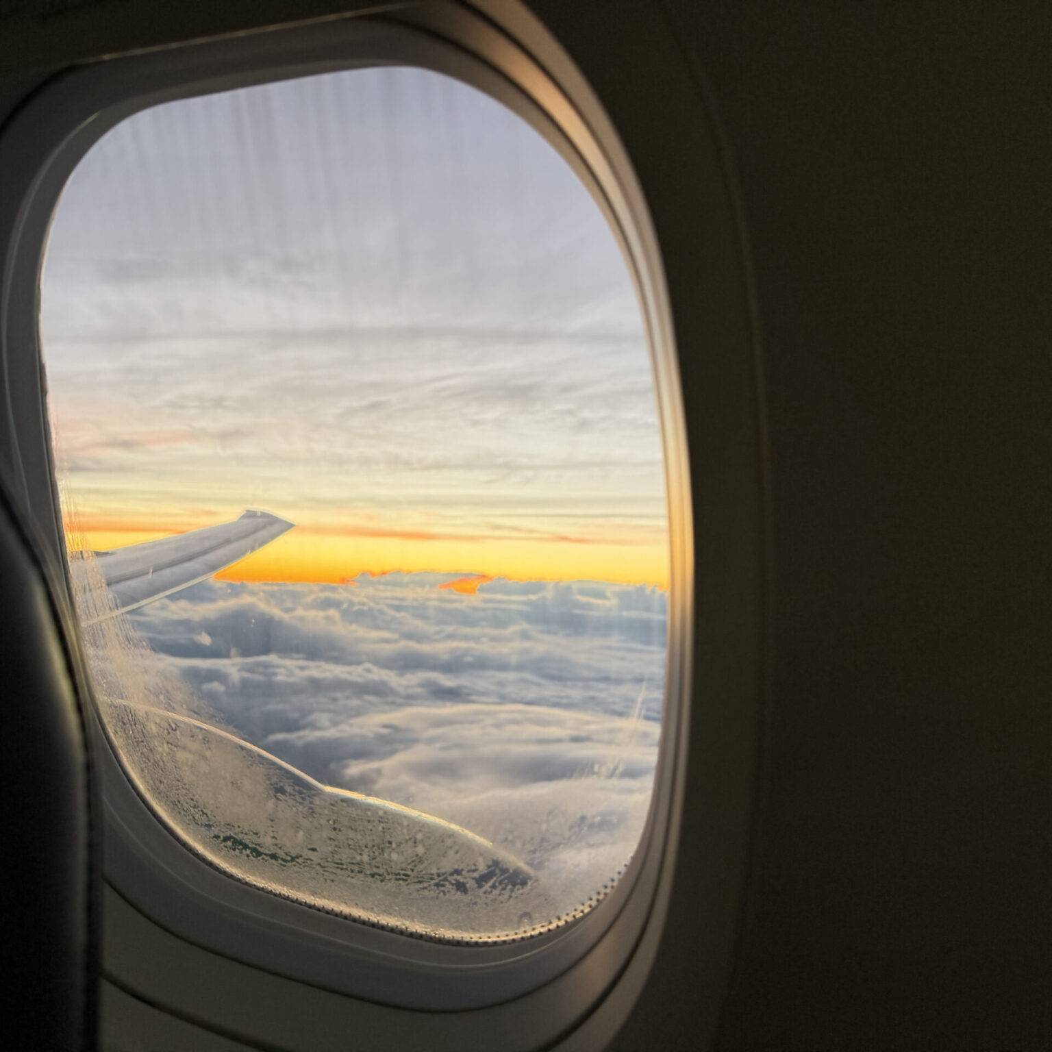 Blick aus einem Flugzeugfenster auf Wolken und einen Sonnenuntergang mit dem Flugzeugflügel im Bild.