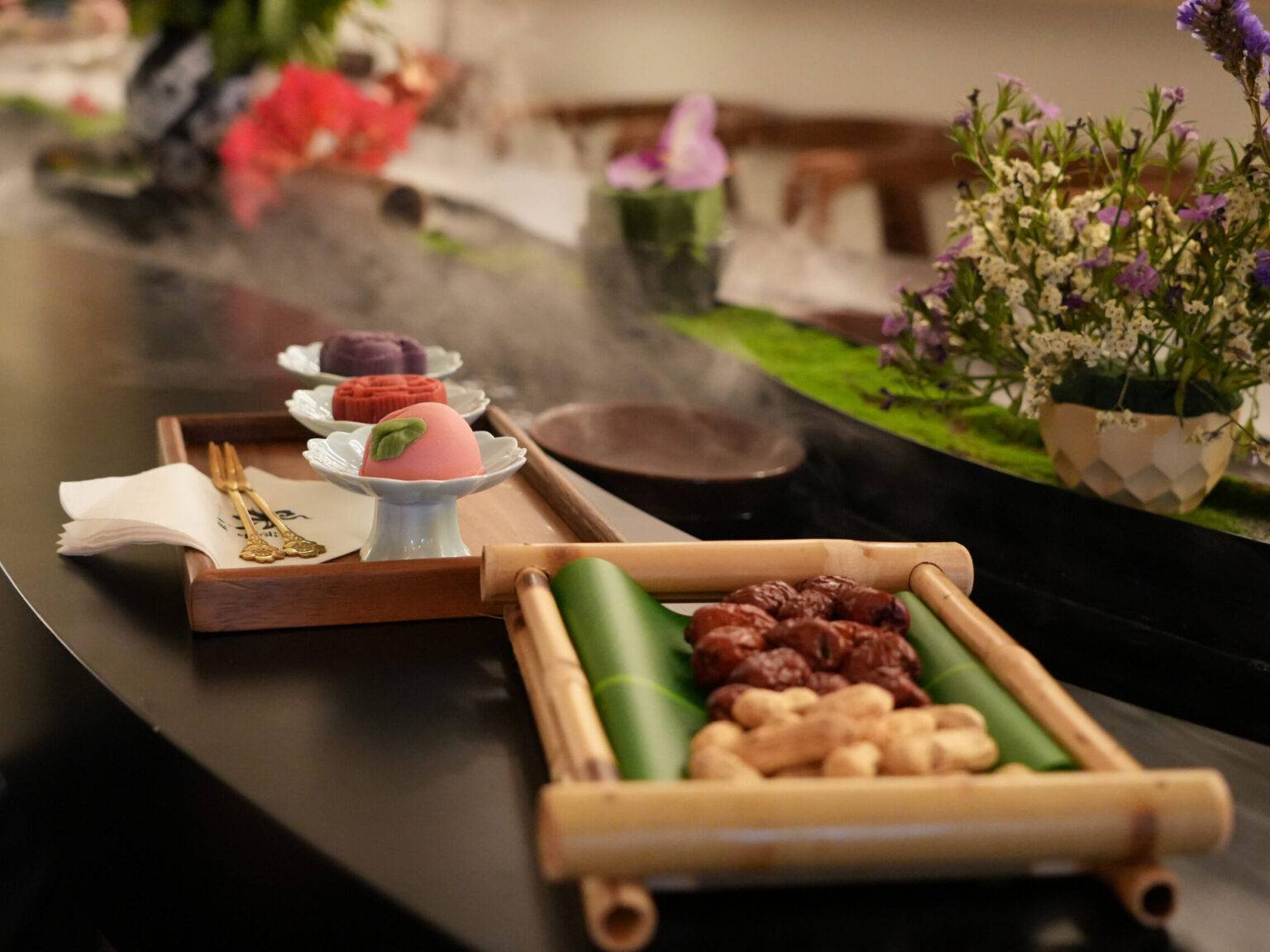 Zwei Tabletts mit verschiedenen Speisen, darunter Desserts und Snacks, auf einem eleganten Tisch mit Blumenarrangements.