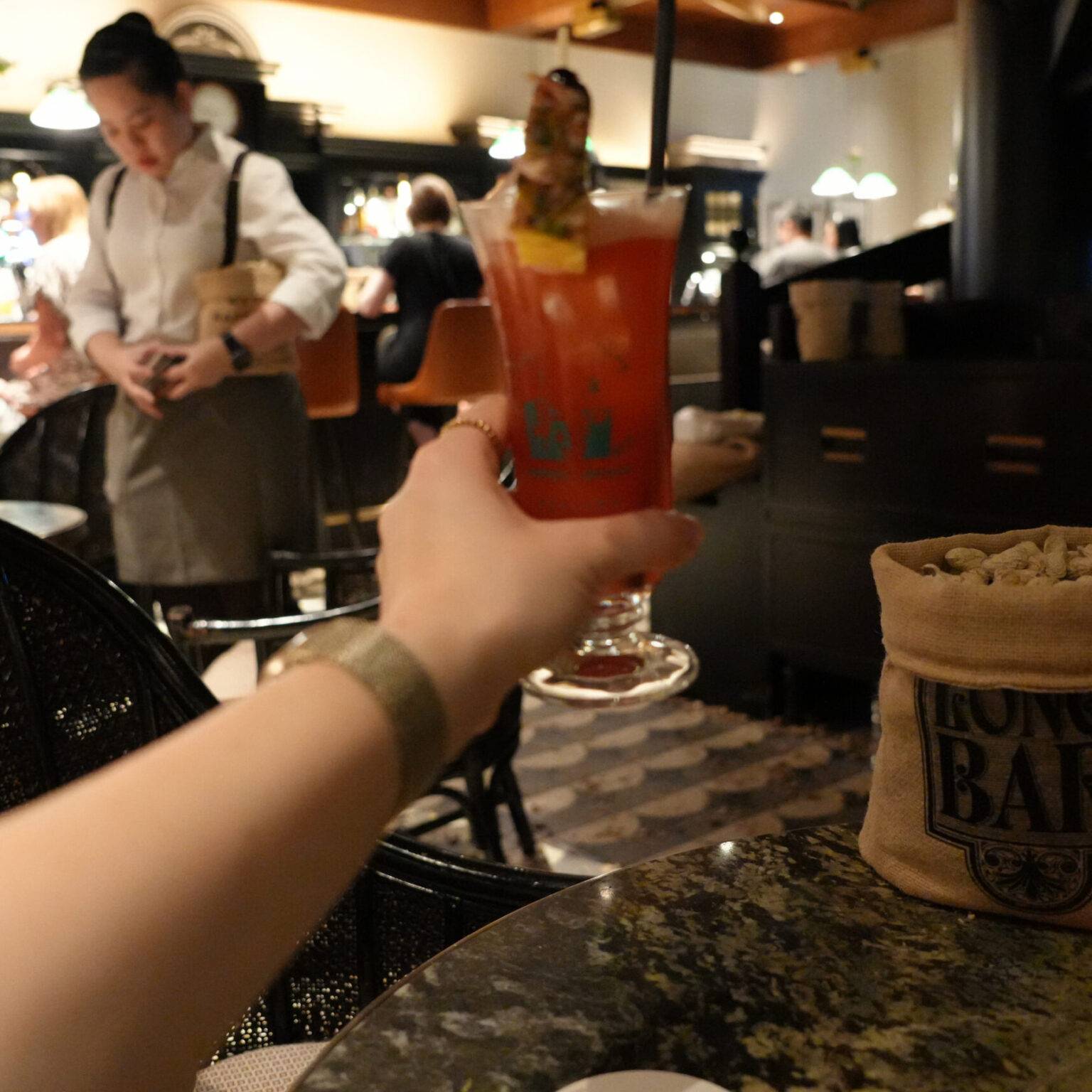 Eine Hand hält einen Singapore Sling Cocktail in der Long Bar, im Hintergrund sitzen Gäste am Tresen