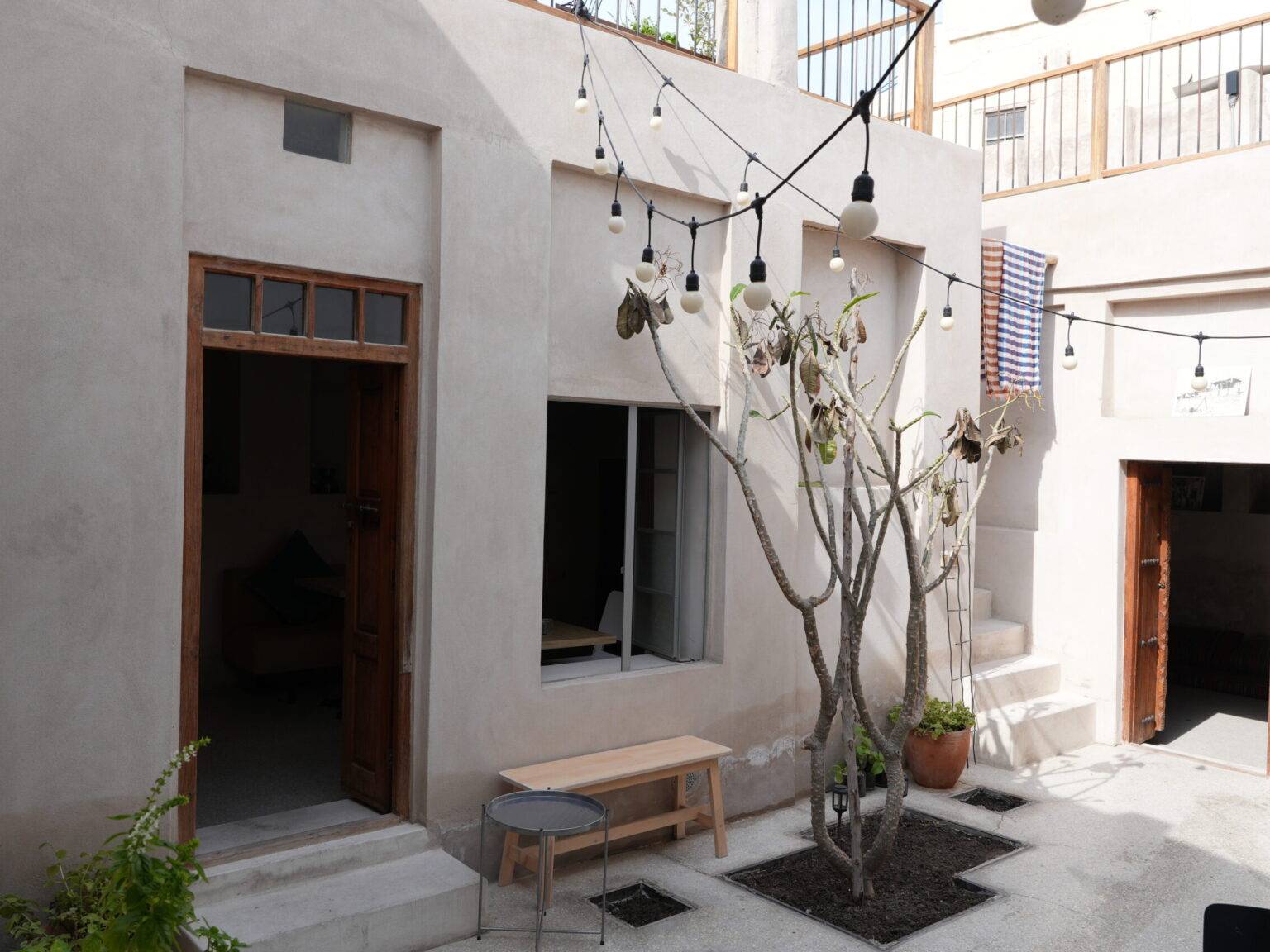 Ruhiger Innenhof des Higher Grounds Café in Muharraq, mit kahlem Baum, Lichterketten und schlichten Holzmöbeln zwischen hellen Steinmauern.