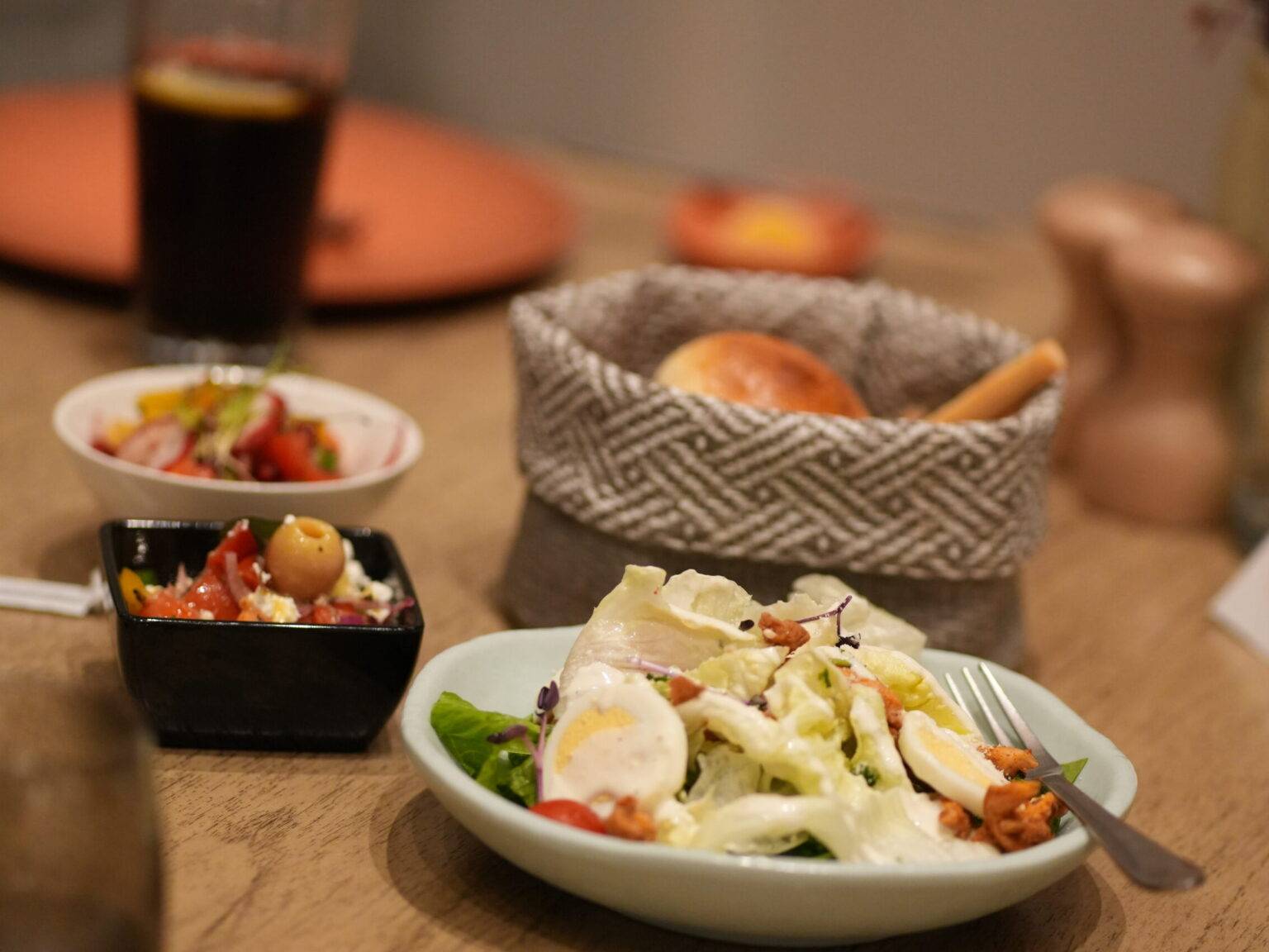 Gedeckter Tisch mit Salat auf weißem Teller, Brotkorb mit frischem Brot und kleinen Schälchen mit Beilagen.