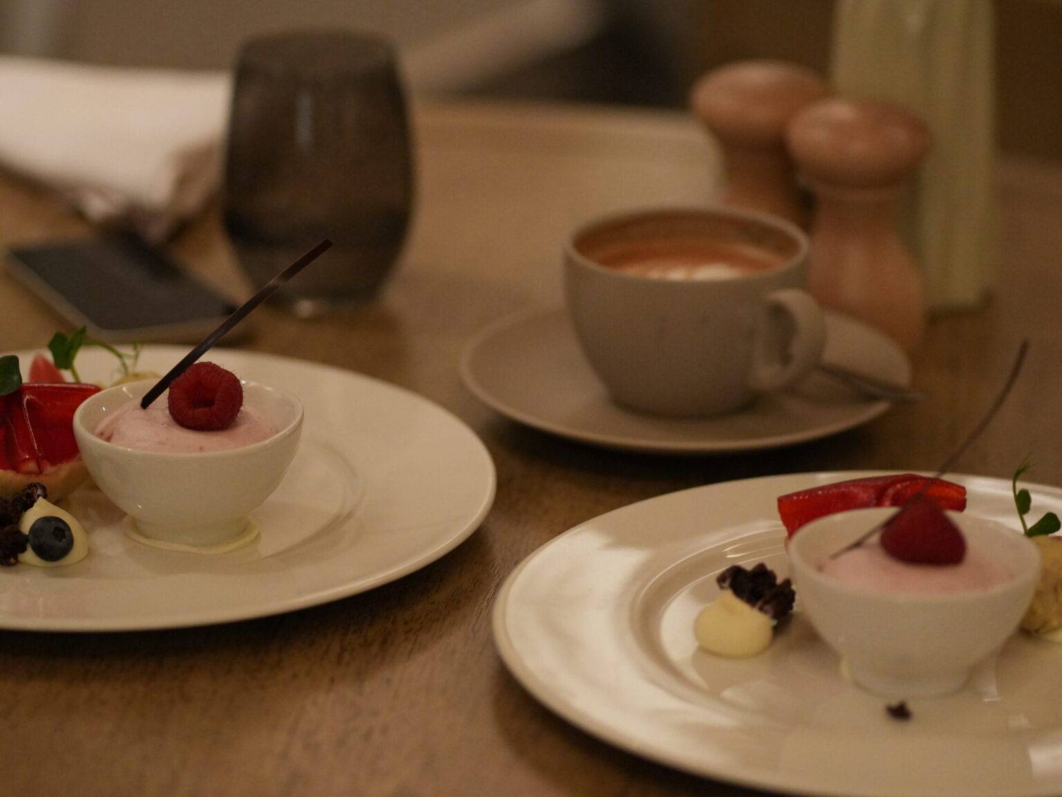 Zwei Dessertteller mit cremiger Süßspeise, garniert mit Beeren und Obst, dazu eine Tasse Cappuccino auf einem Tisch.