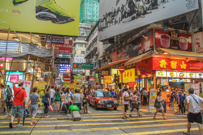 In einer belebten Straße von Hongkong wuseln zwischen den bunten Shops Menschen und Fahrzeuge.