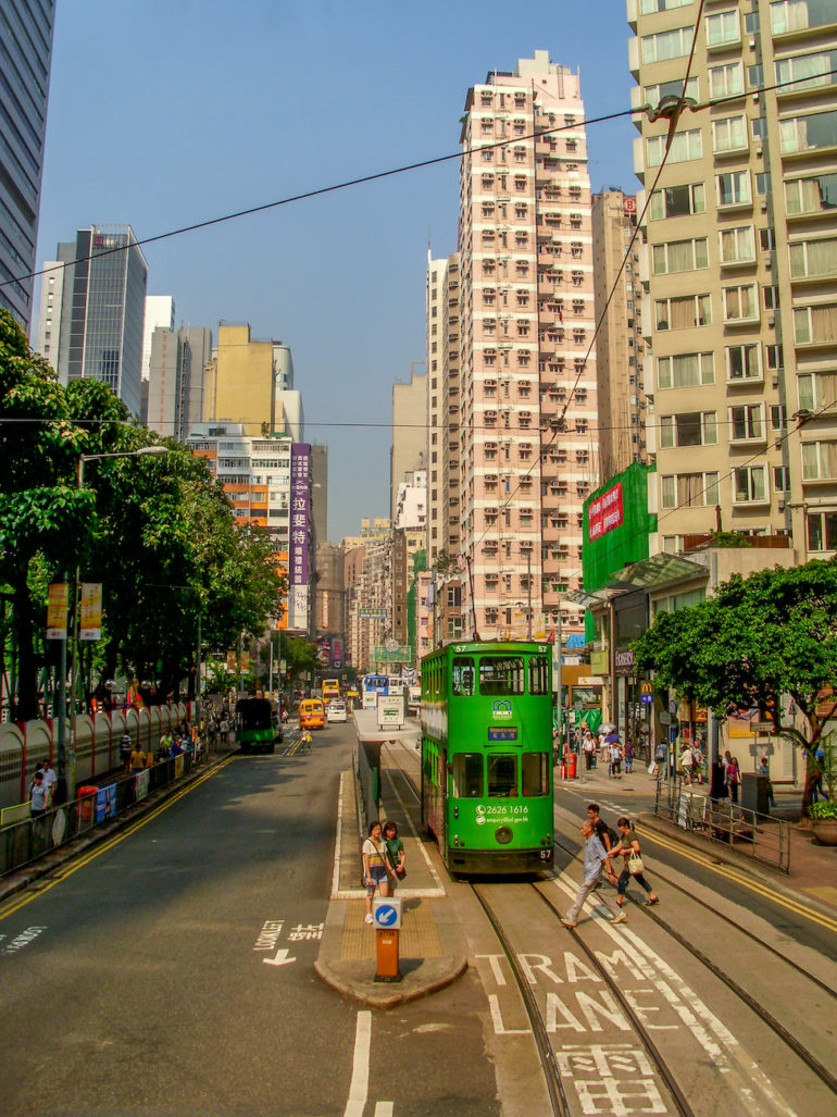 Ein historischen Straßenbahn in grün fährt eine Straße von Hongkong entlang vorbei an Hochhäusern.