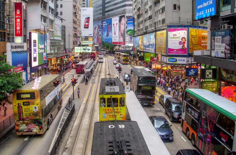 In Hongkongs Viertel Causeway Bay tummeln sich Fahrzeuge, Busse, Straßenbahnen und Menschen entlang der Straßen mit Läden und bunt plakatierten Hausfassaden.
